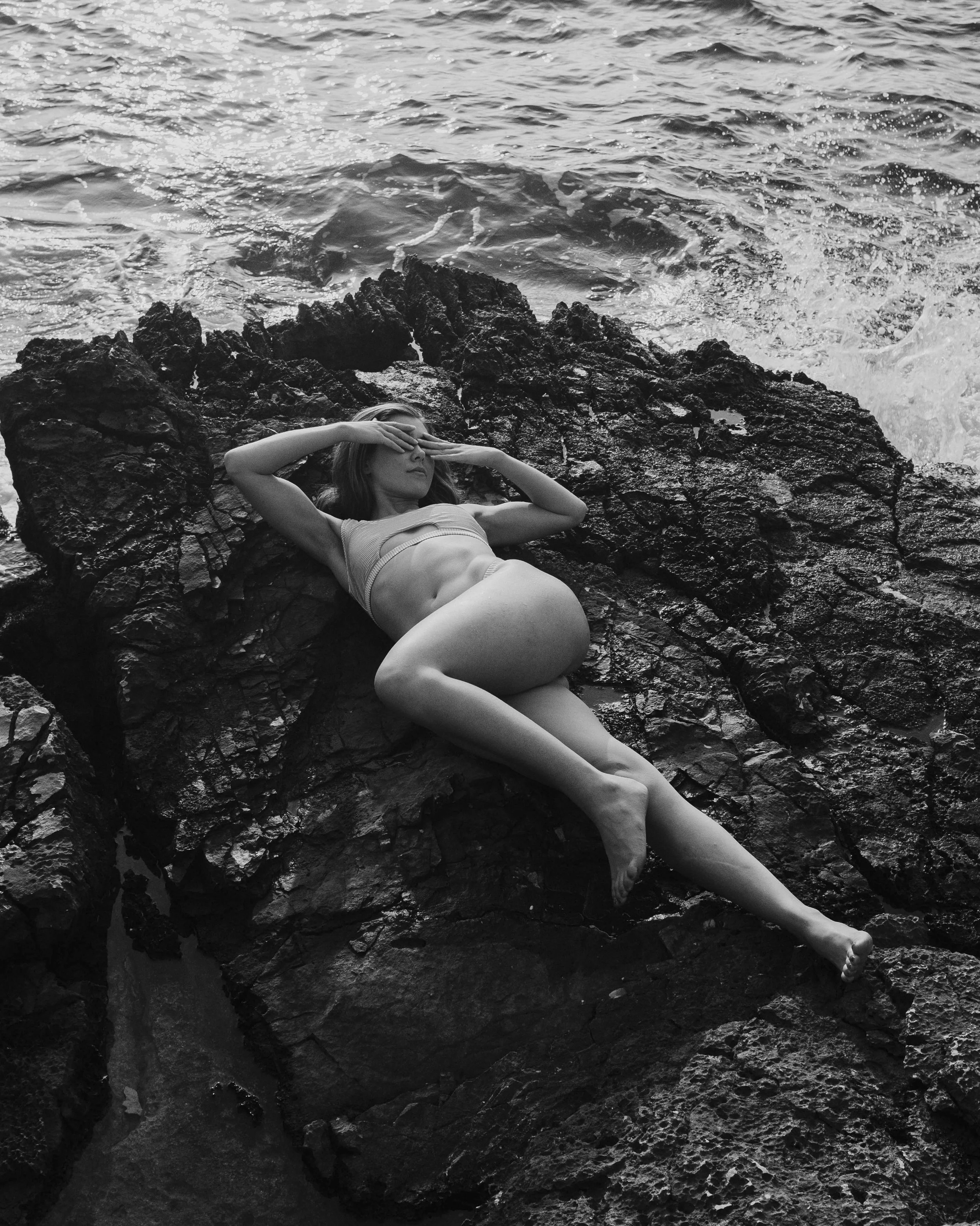 A woman lying on dark rocks by the water, wearing a swimsuit, with one arm covering her eyes and the other resting on her forehead.
