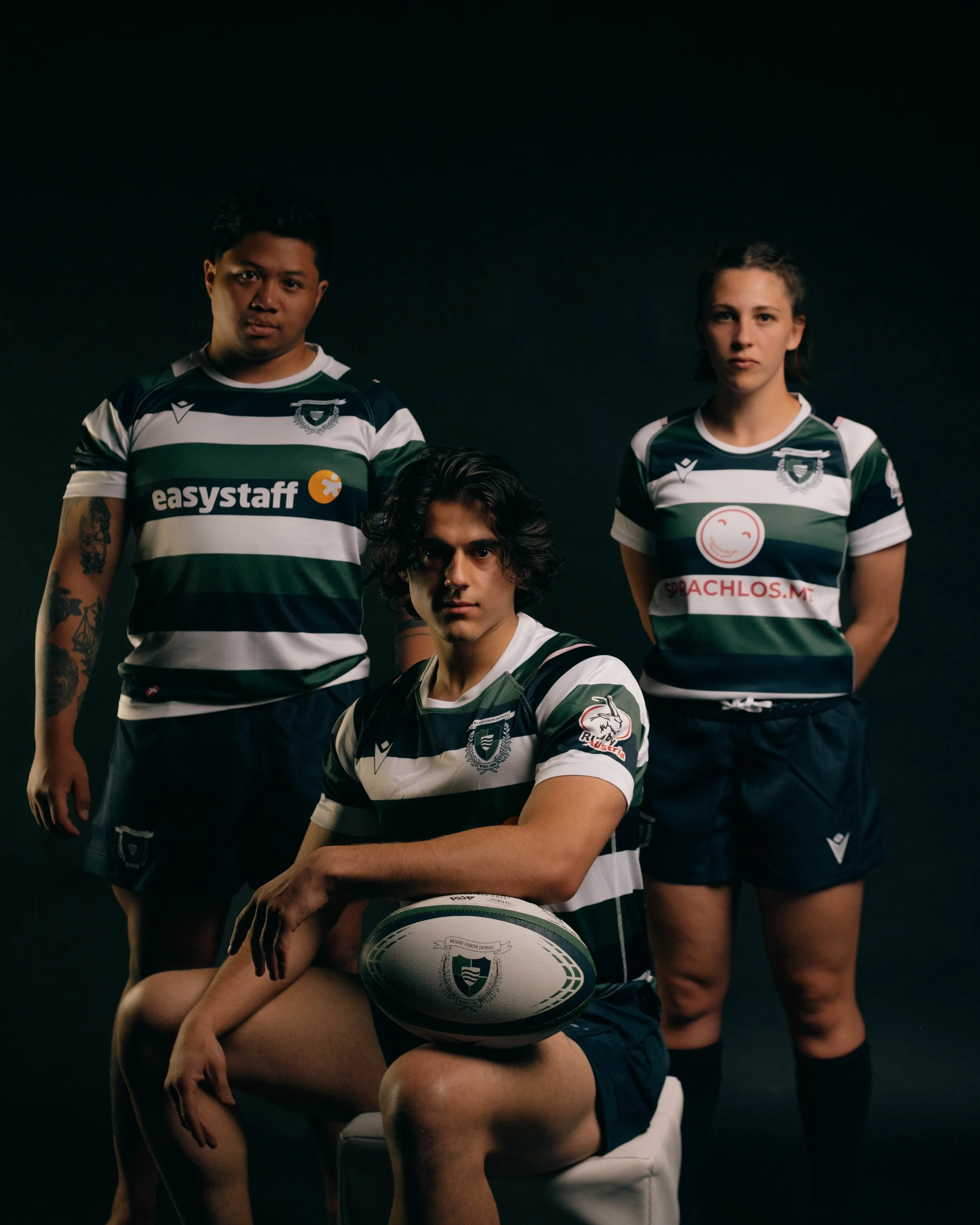 Three rugby players in green and white striped jerseys posing against a dark background. One player is seated with a rugby ball, and two are standing behind him.