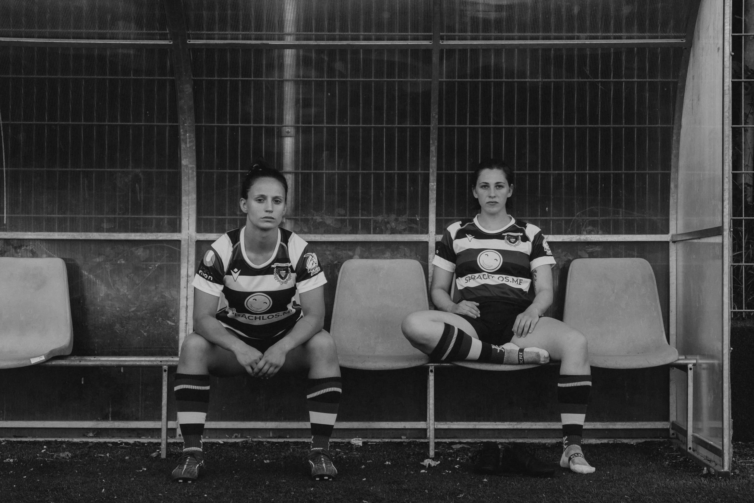 Two female soccer players sitting on a bench in a sports dugout, both wearing striped jerseys, shorts, and socks, with serious expressions, in black and white.