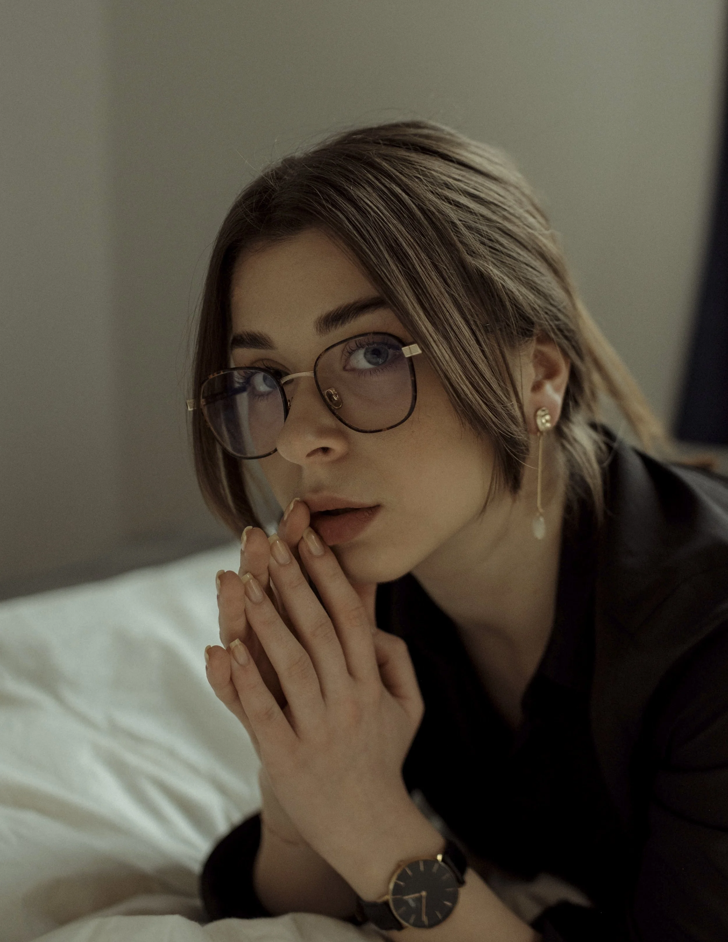 Close-up of a young woman with short brown hair, wearing glasses, a black shirt, and gold jewelry, sitting on a bed with a neutral-colored wall in the background.