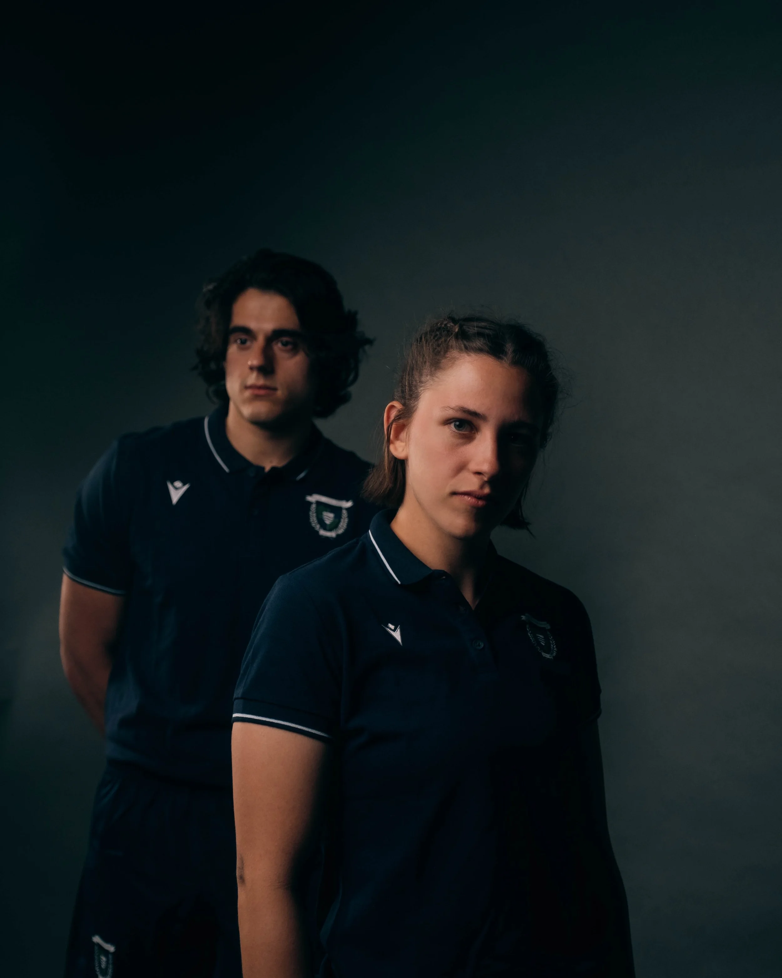 A young woman and man wearing matching navy blue athletic shirts with logos, standing against a dark gray background. The woman is in the foreground, and the man is slightly behind her.