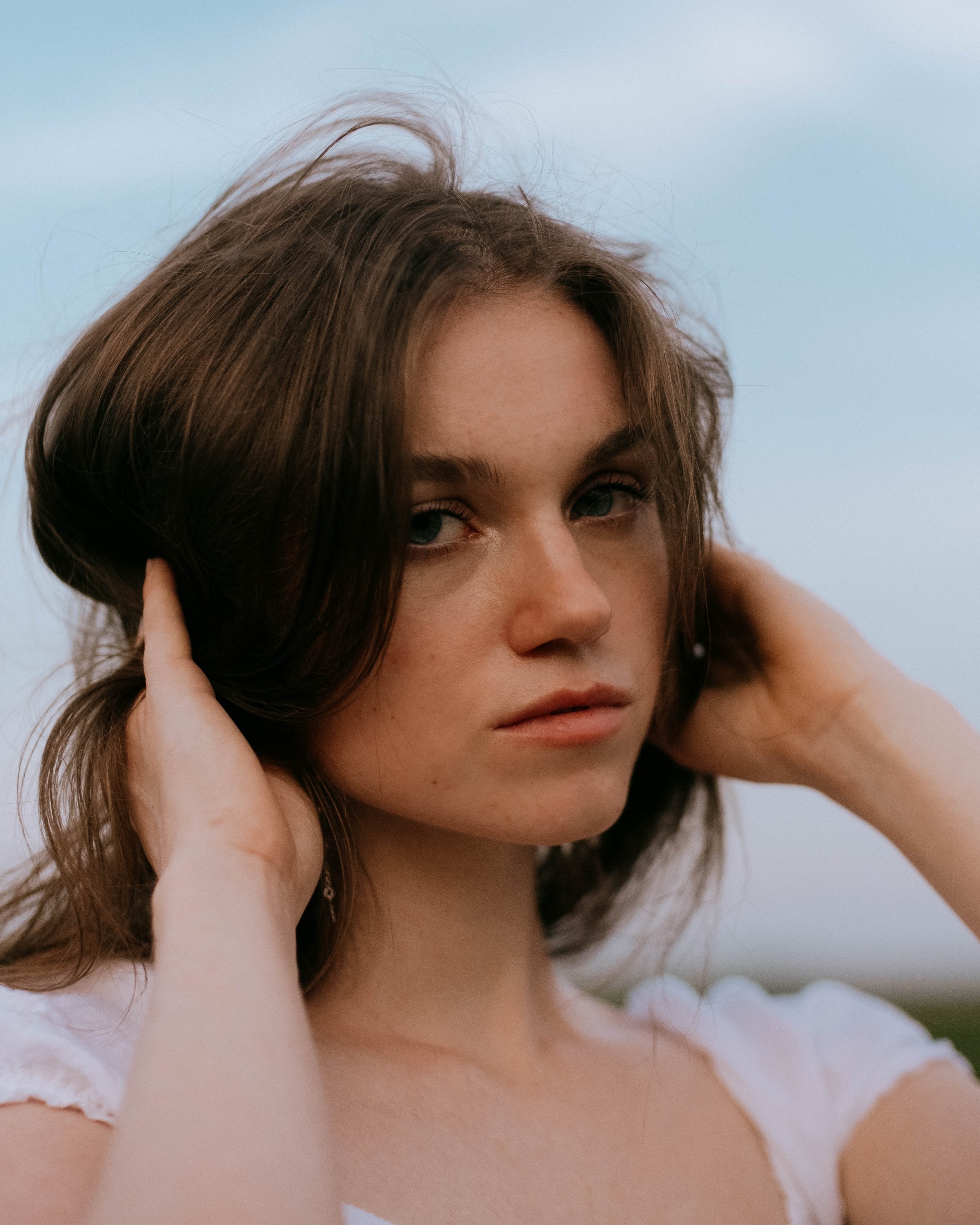 Close-up of a woman with brown hair, blue eyes, and a neutral expression, touching her hair with both hands, outdoors with a cloudy sky background.