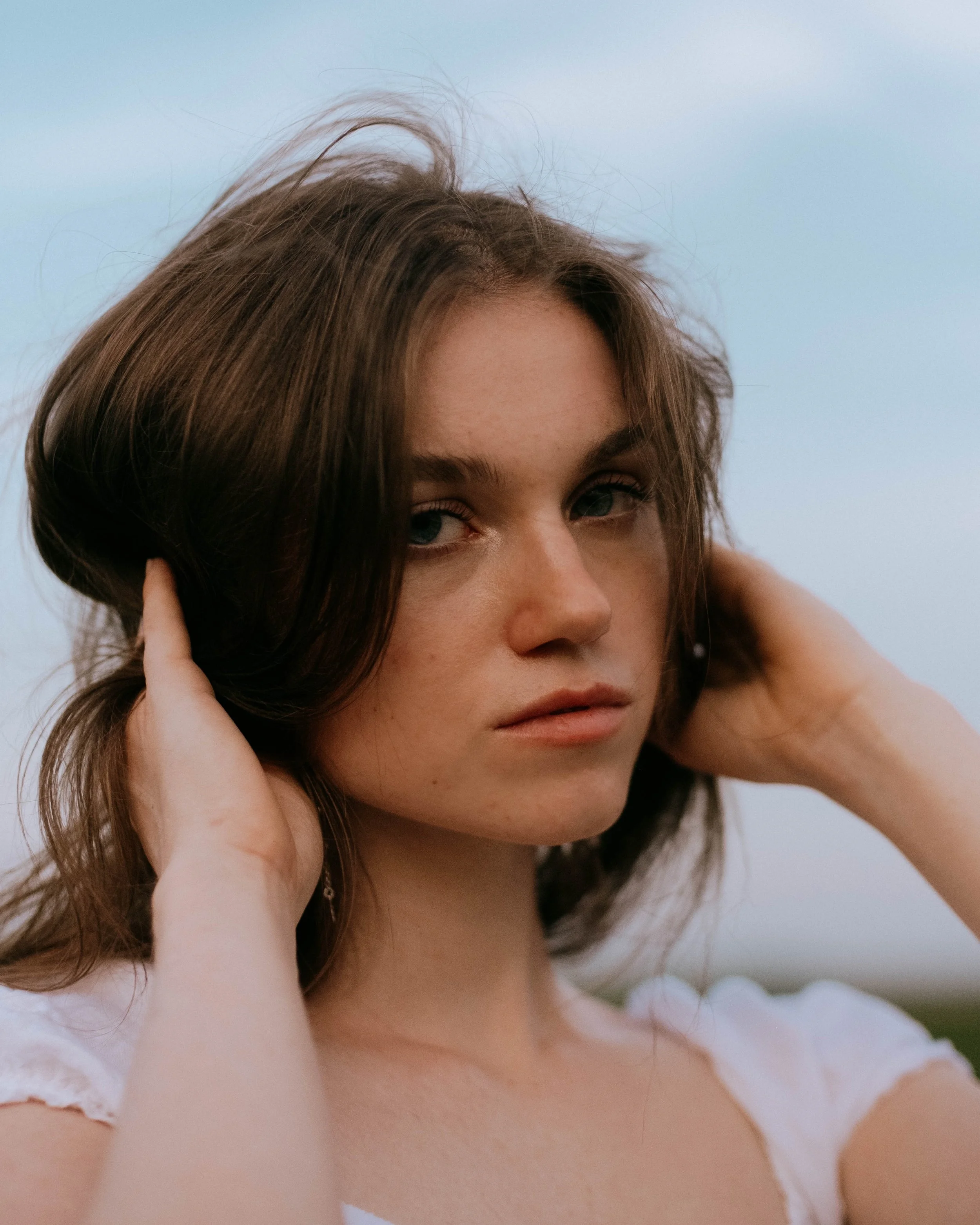 Close-up of a woman with brown hair, blue eyes, and a neutral expression, touching her hair with both hands, outdoors with a cloudy sky background.