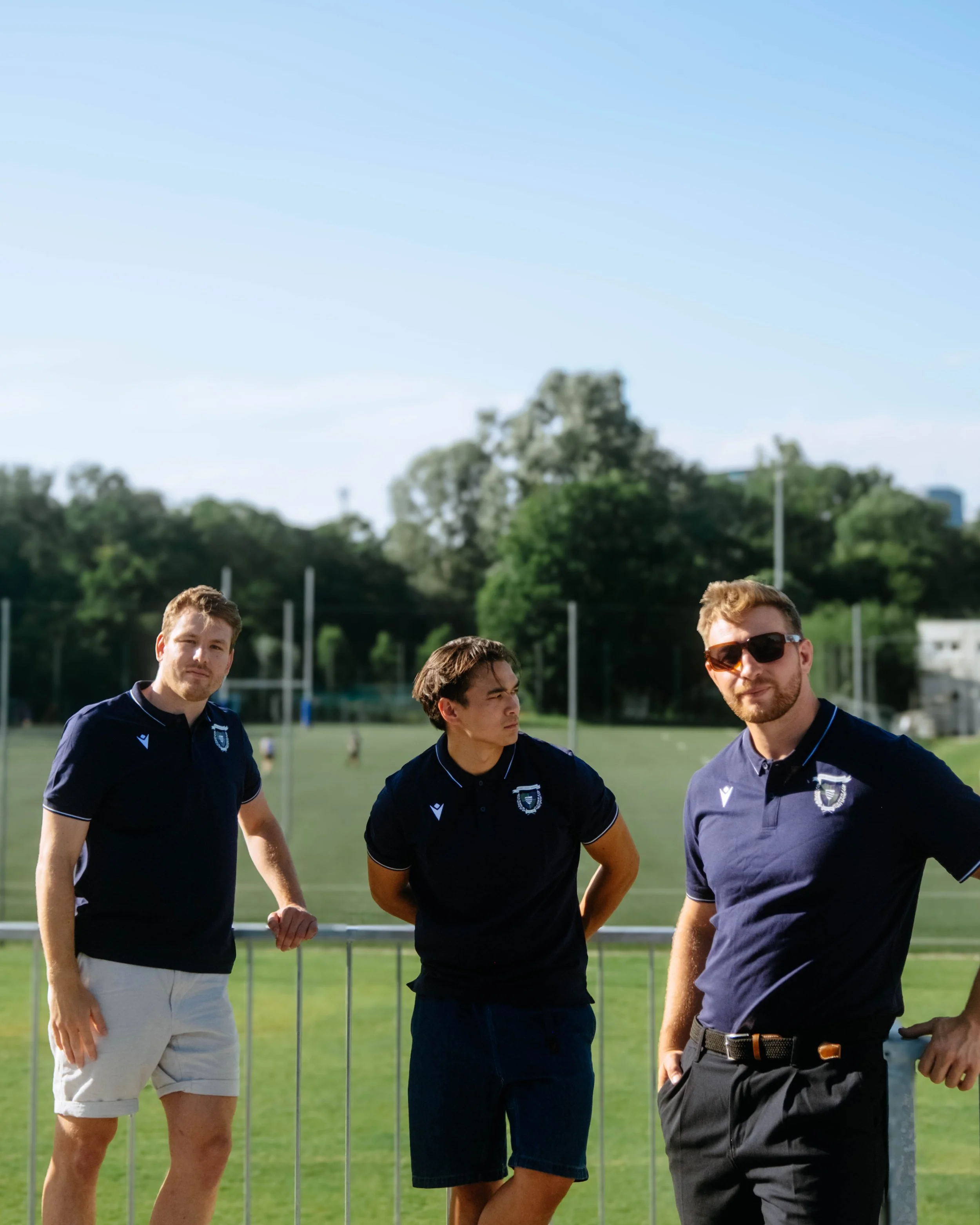 Three young men standing outdoors on a sports field, wearing navy blue polo shirts with a crest, with trees and a blue sky in the background.