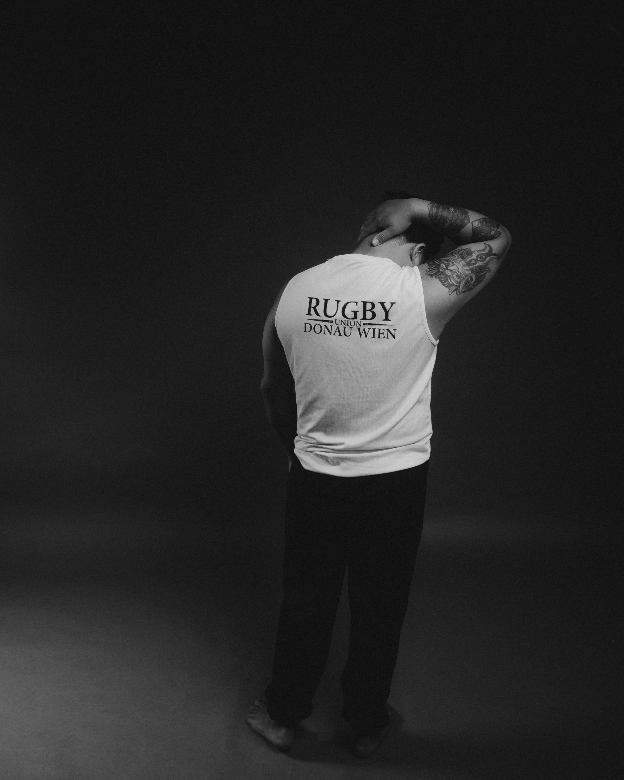 A person standing on a dark background, facing away, wearing a sleeveless shirt with "RUGBY UNION DONAU WIEN" printed on the back, with tattoos on their arms and head tilted down with one hand on their head.