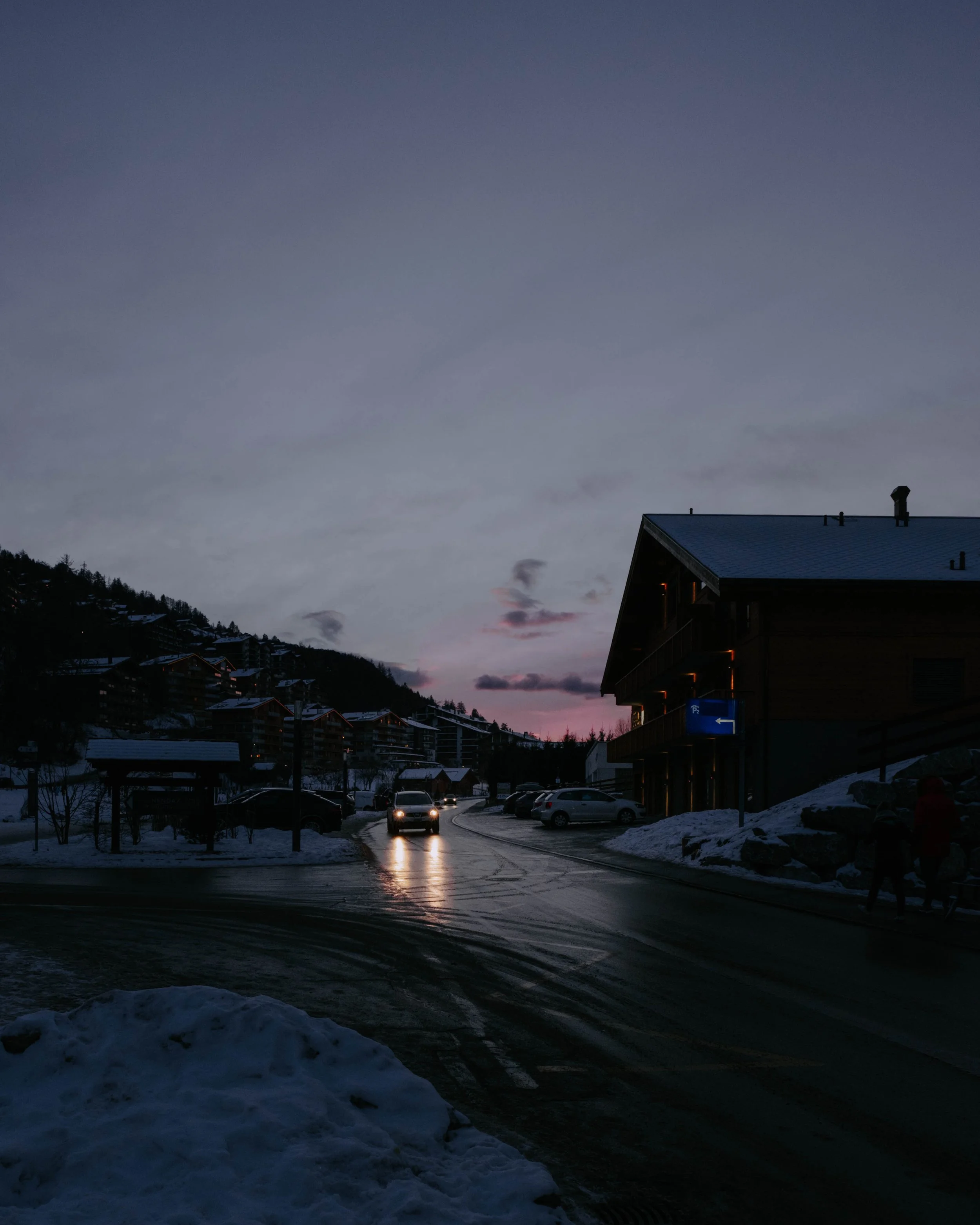 A snowy parking lot at dusk with several cars, a modern building on the right, mountains in the background, and pedestrians walking on the sidewalk.