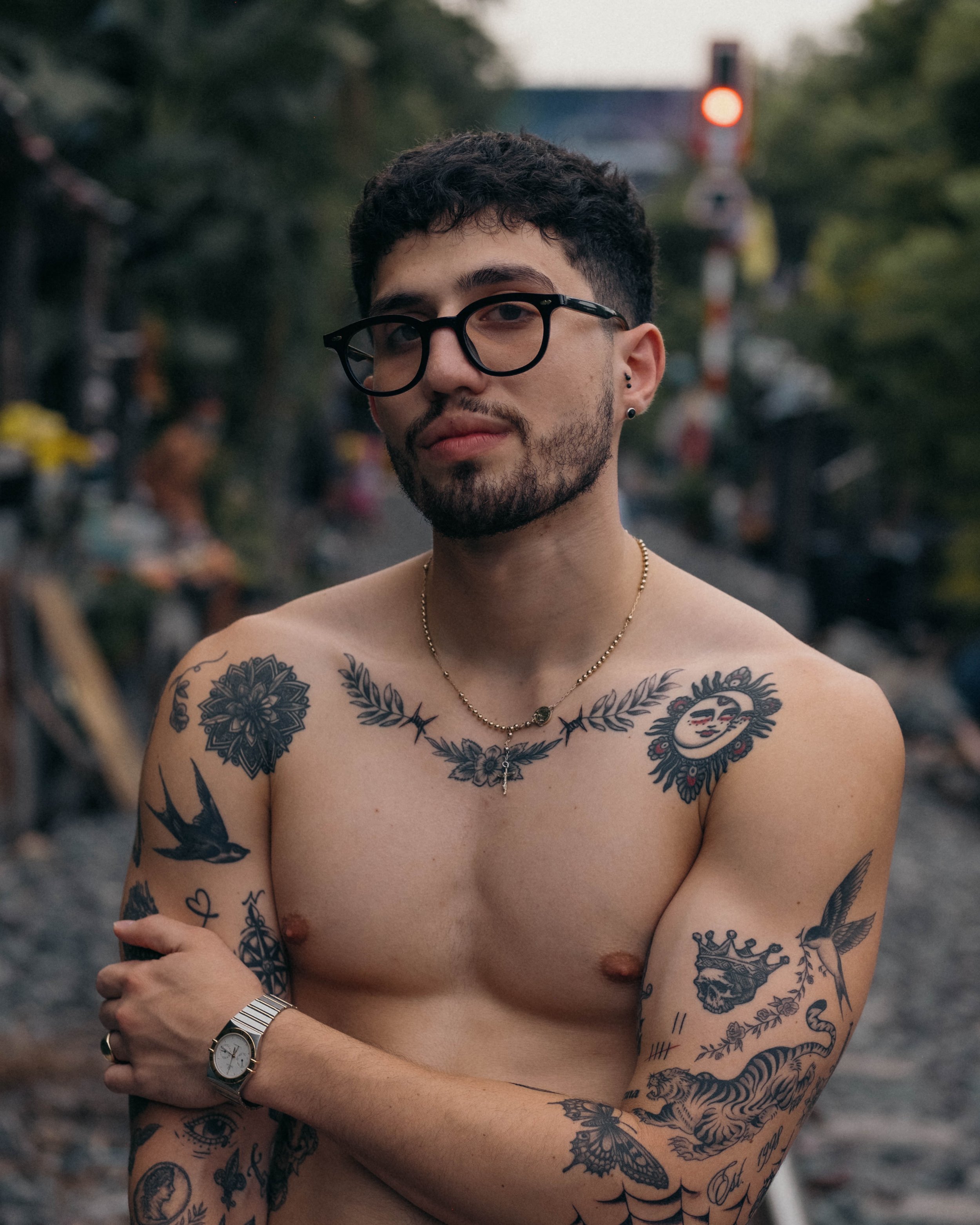 A shirtless young man with glasses, tattoos on his arms and chest, wearing a watch, standing outdoors with a blurred background of trees and a railway crossing.
