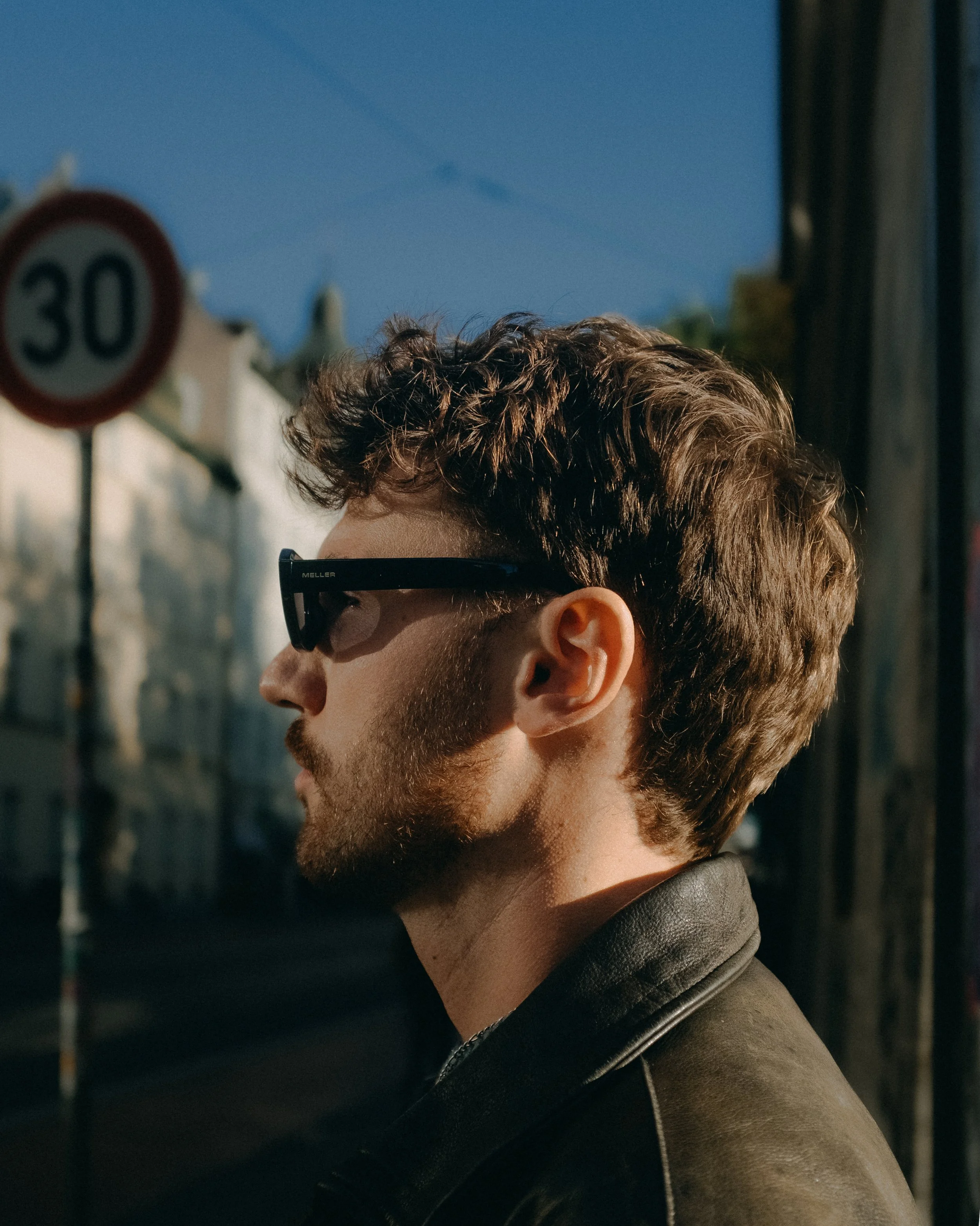 Profile of a man with curly brown hair, beard, wearing sunglasses and a leather jacket, standing outdoors near a pole with a '30' speed limit sign, with a clear blue sky in the background.