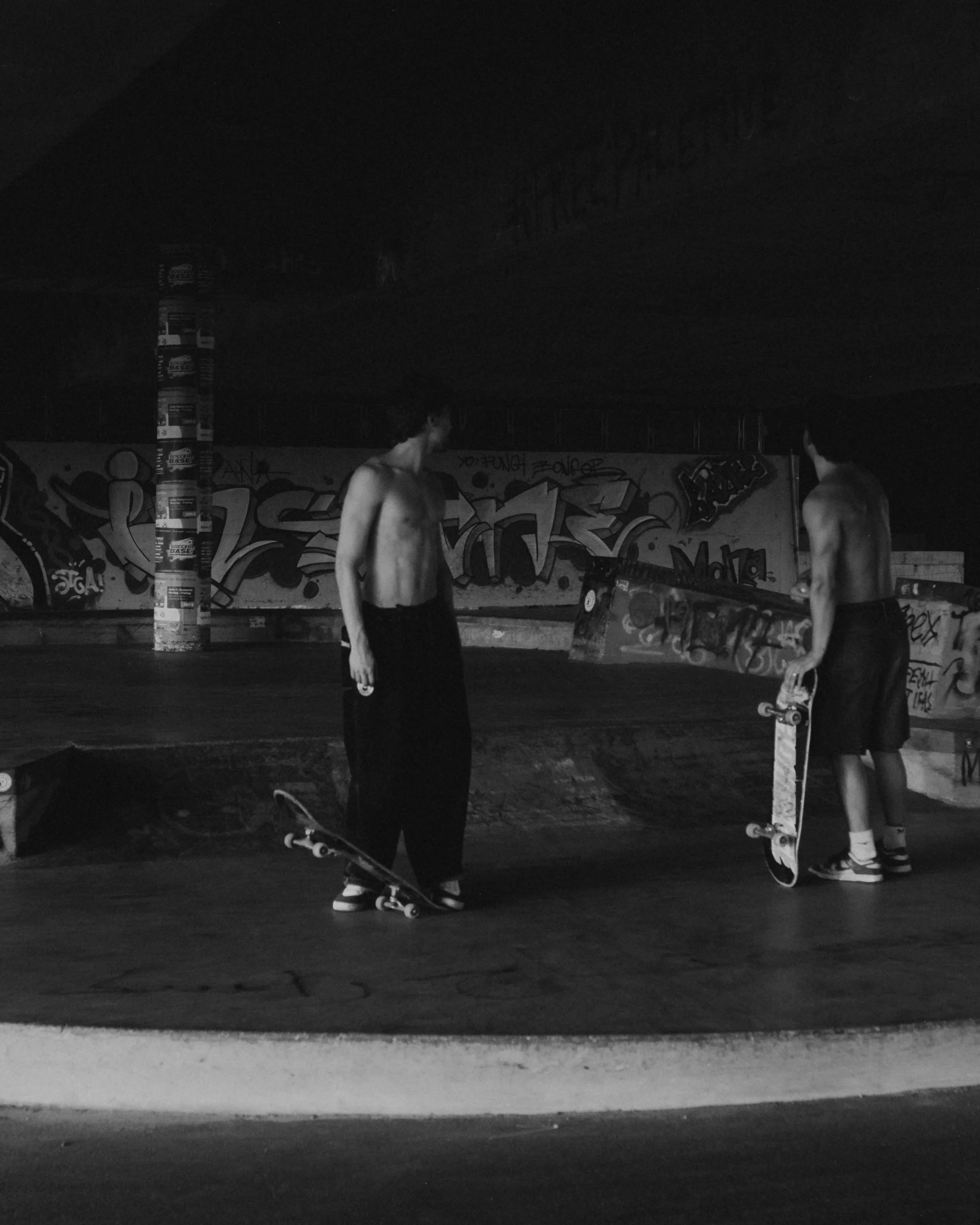 Two shirtless young men with skateboards in an indoor skate park with graffiti on the walls, black and white photo.