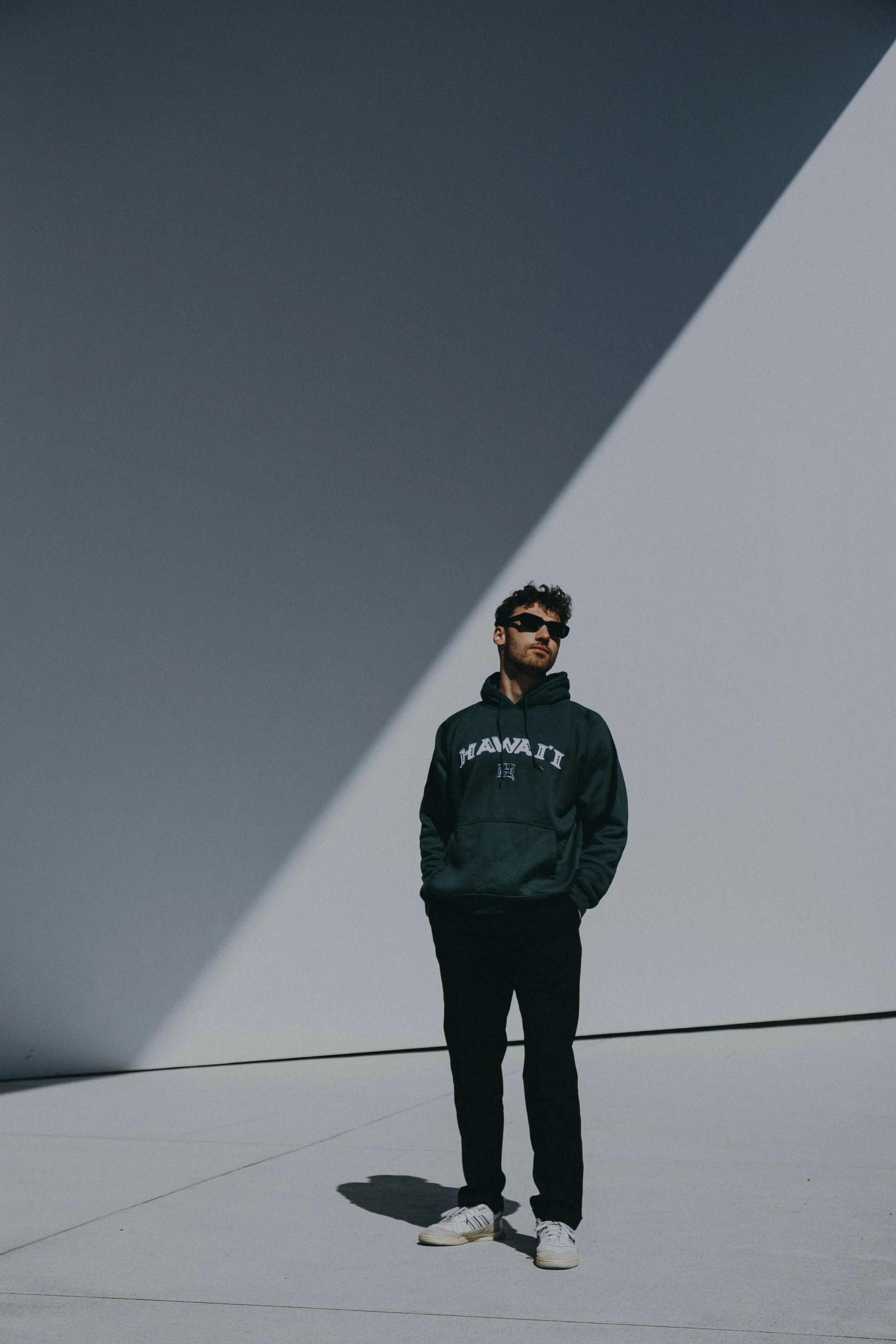 A man in dark sunglasses, a dark hoodie with 'Hawaii' written on it, black pants, and white sneakers standing outdoors against a minimalist background of large, geometric concrete or architectural elements with shadows.