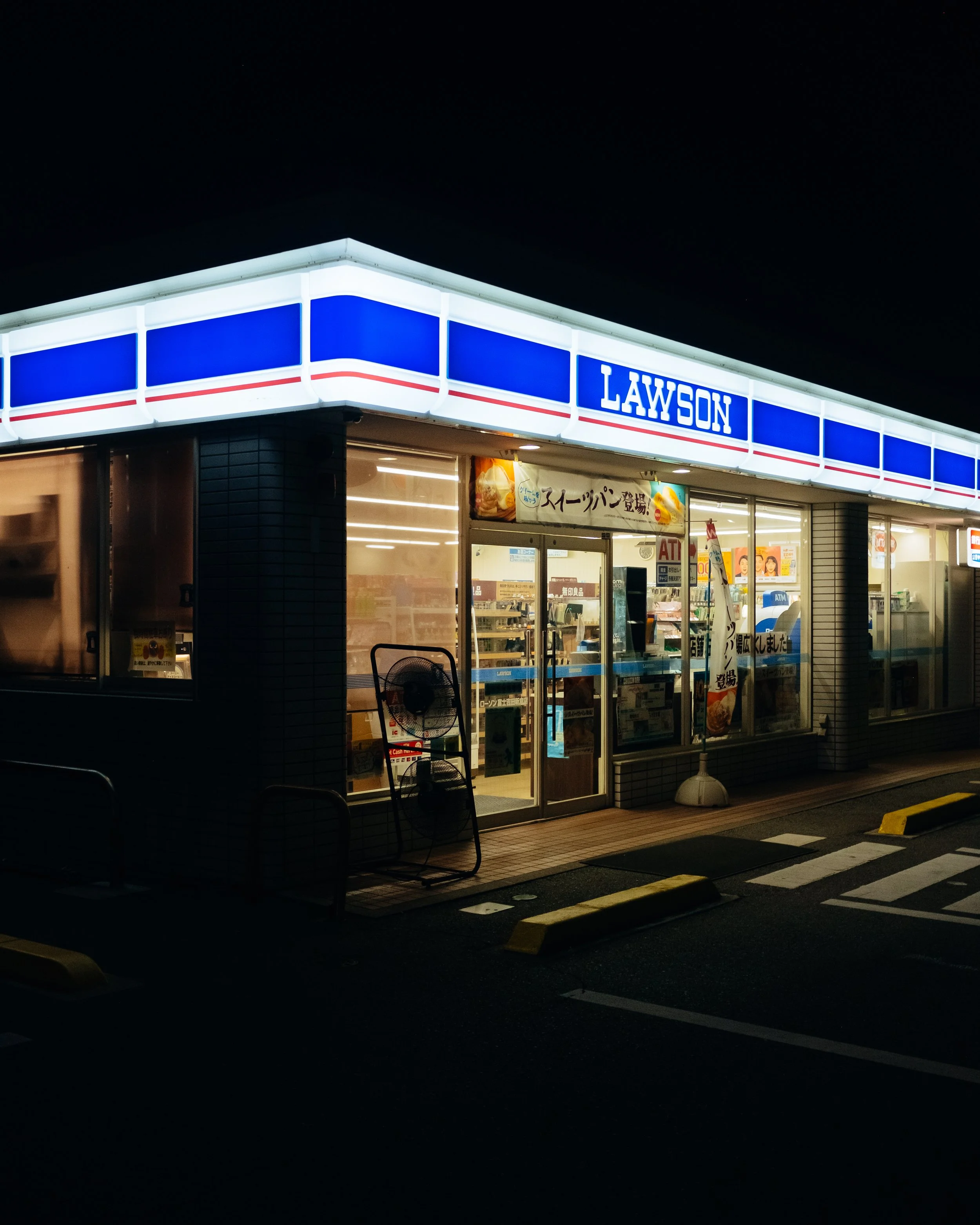 Night view of a Lawson convenience store with illuminated signage, glass doors, and a parking lot in the foreground.