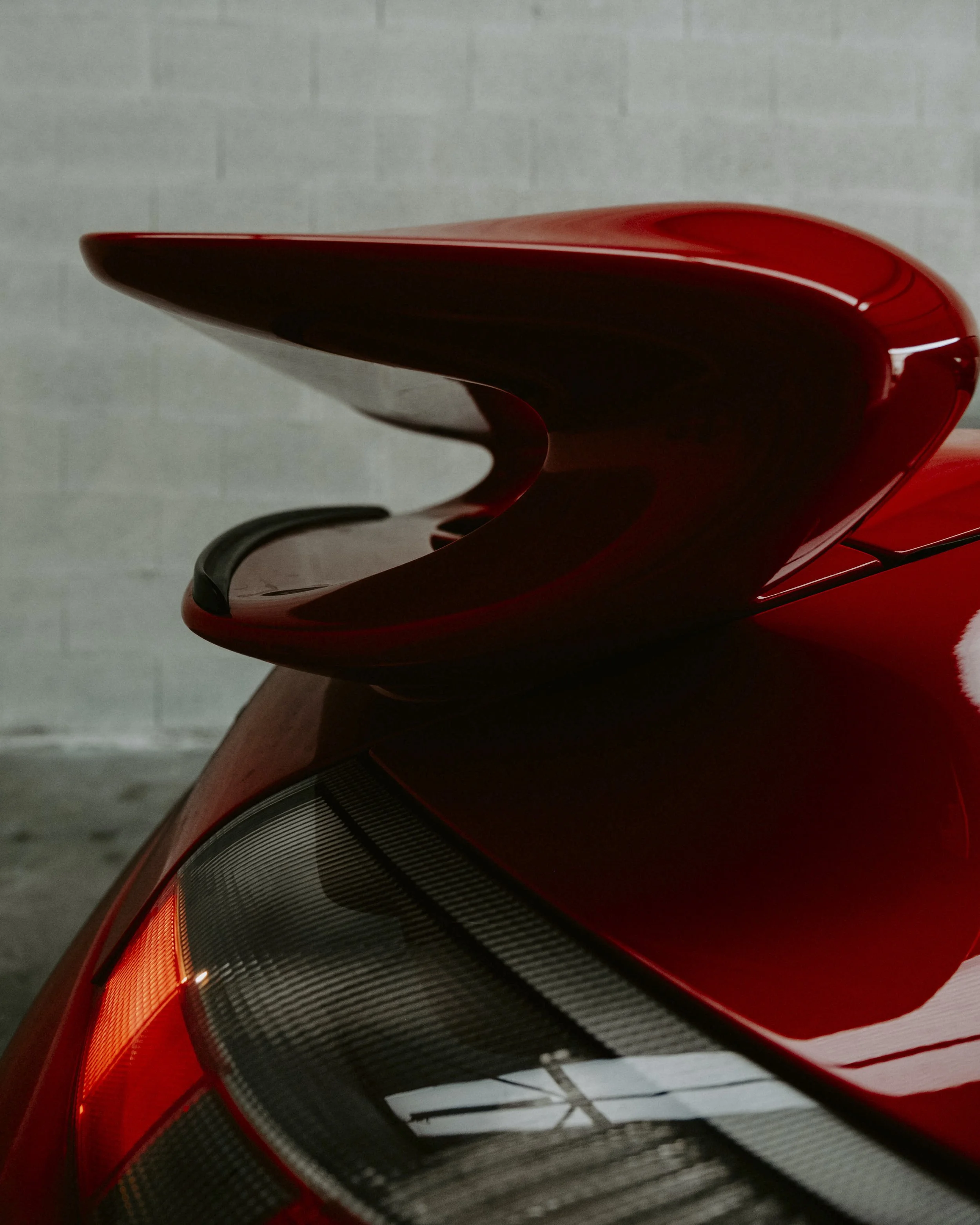 Close-up of a red sports car with a prominent rear spoiler, showing the tail light and part of the car's body against a plain wall.