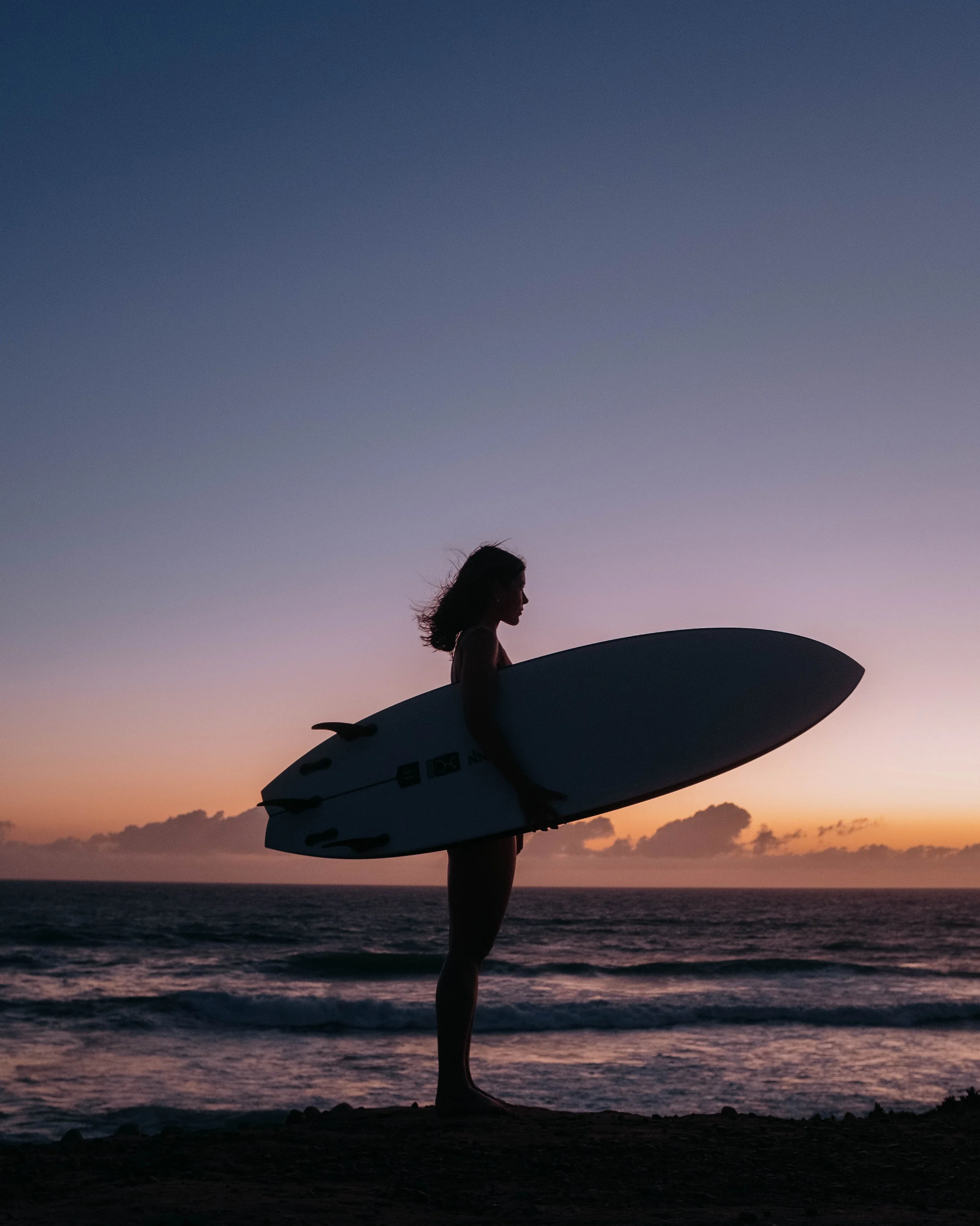 Silhouette of a woman holding a surfboard on a beach during sunset with colorful sky and ocean waves.