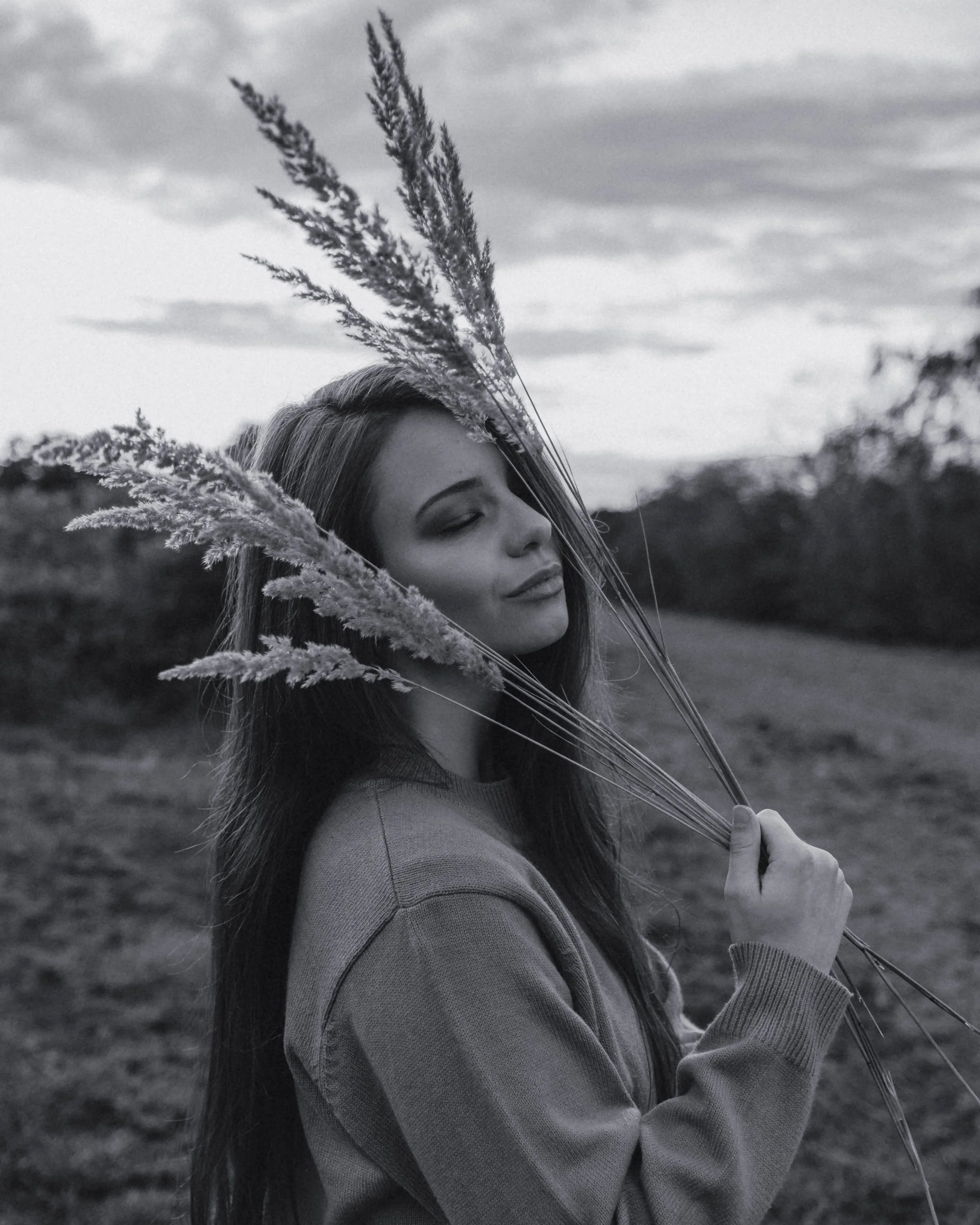 A woman with long hair holding a bunch of tall grass, standing outdoors with a cloudy sky in the background.