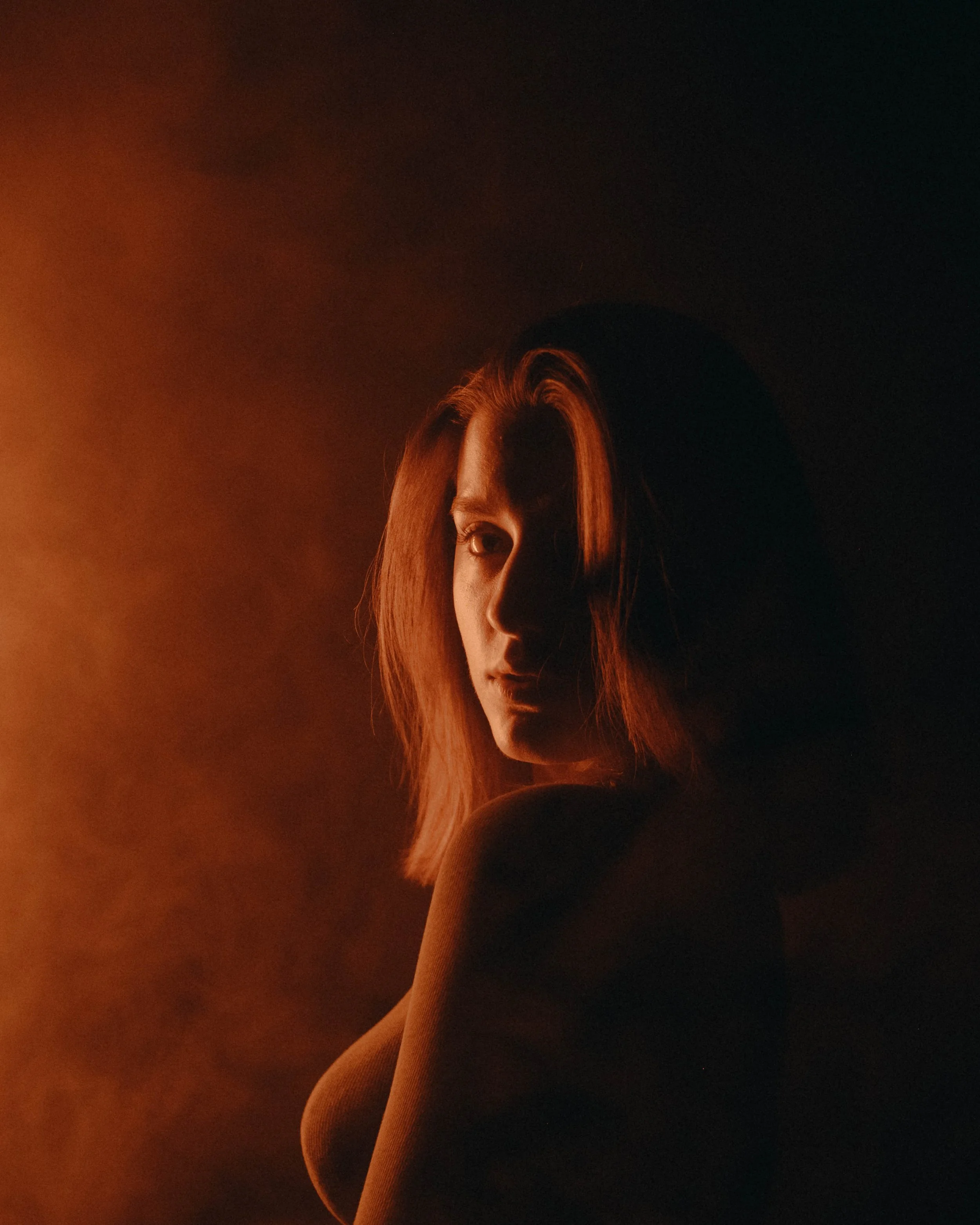 A woman with medium-length hair looking to the camera, partially illuminated by warm orange light on her face, with dark shadow on the other side, against a dark background.
