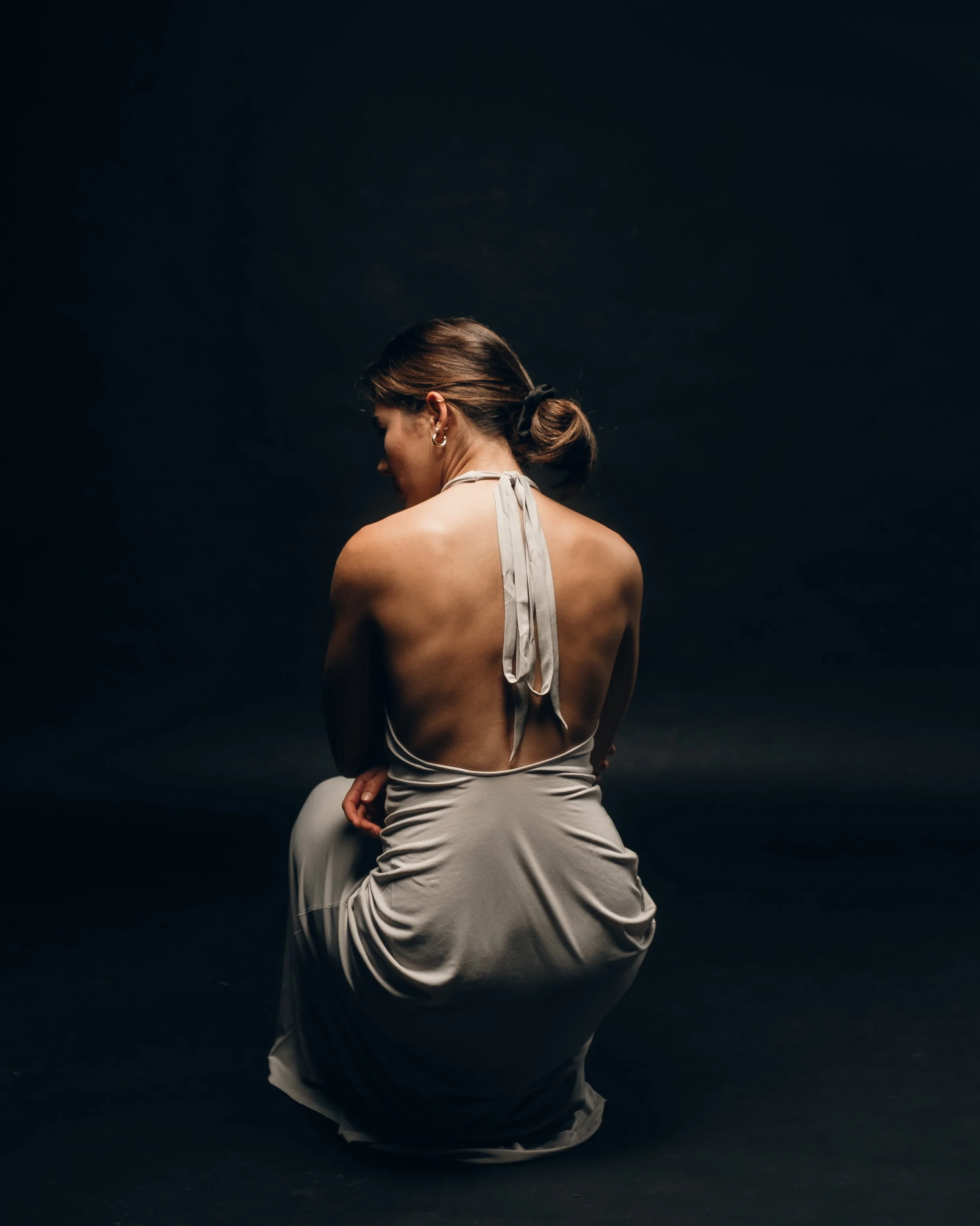 A woman in a light-colored dress kneeling in front of a dark background, with her back facing the camera, her head turned to the side, and her hair tied back.