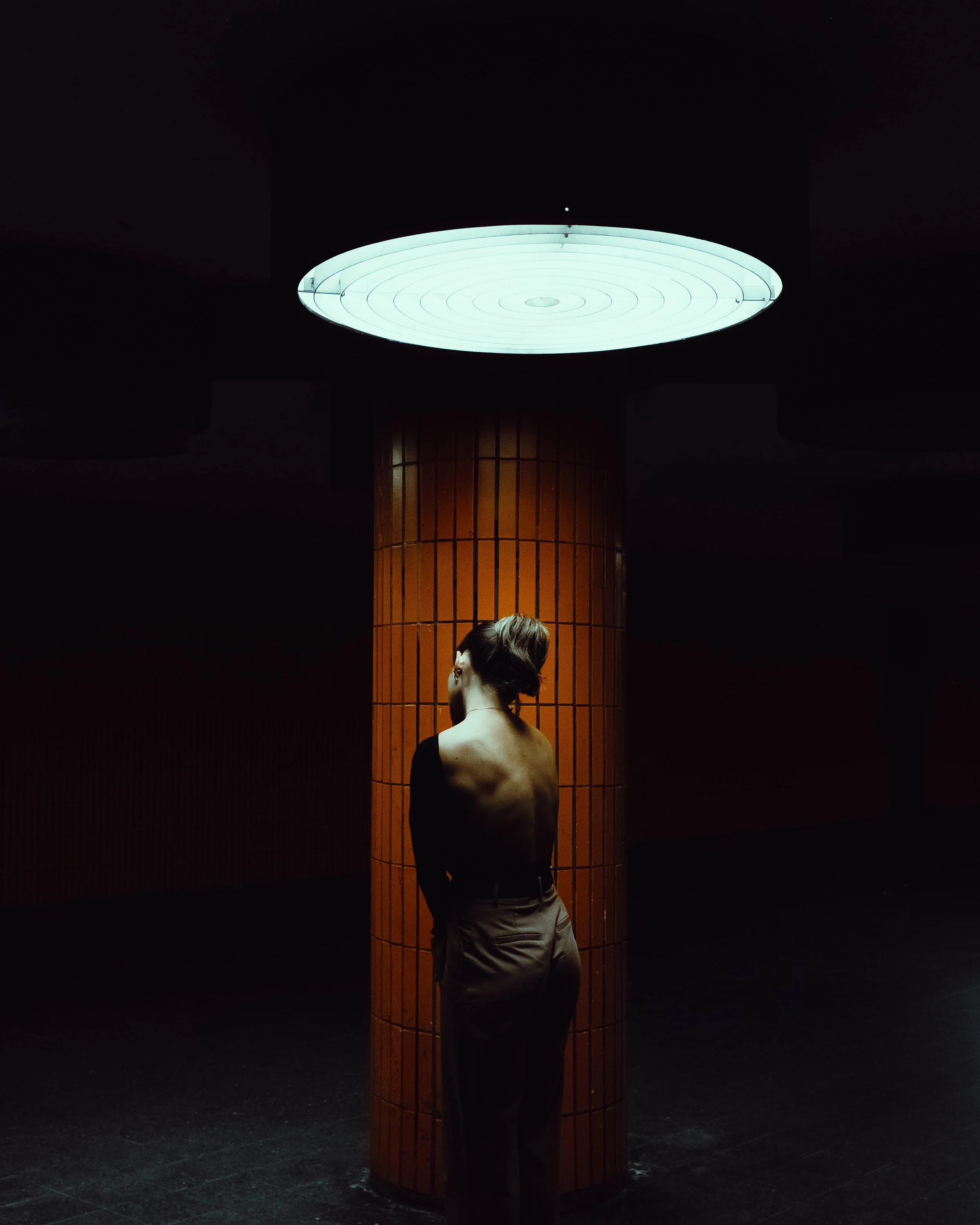 A woman with short hair, wearing a black top and beige pants, standing with her back to the camera under a large, glowing circular ceiling light in a dimly lit space.
