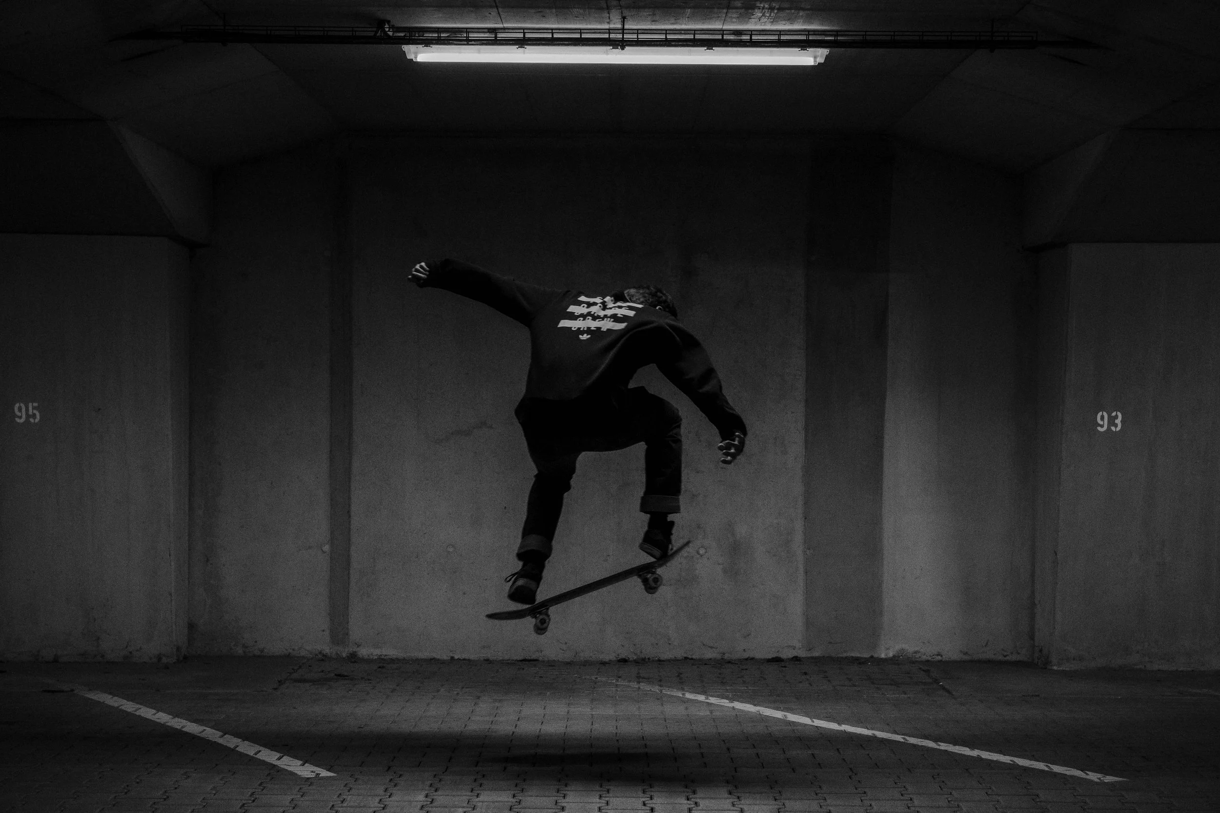A skateboarder in a dark hoodie performs a jump in a dimly lit parking garage with concrete walls.