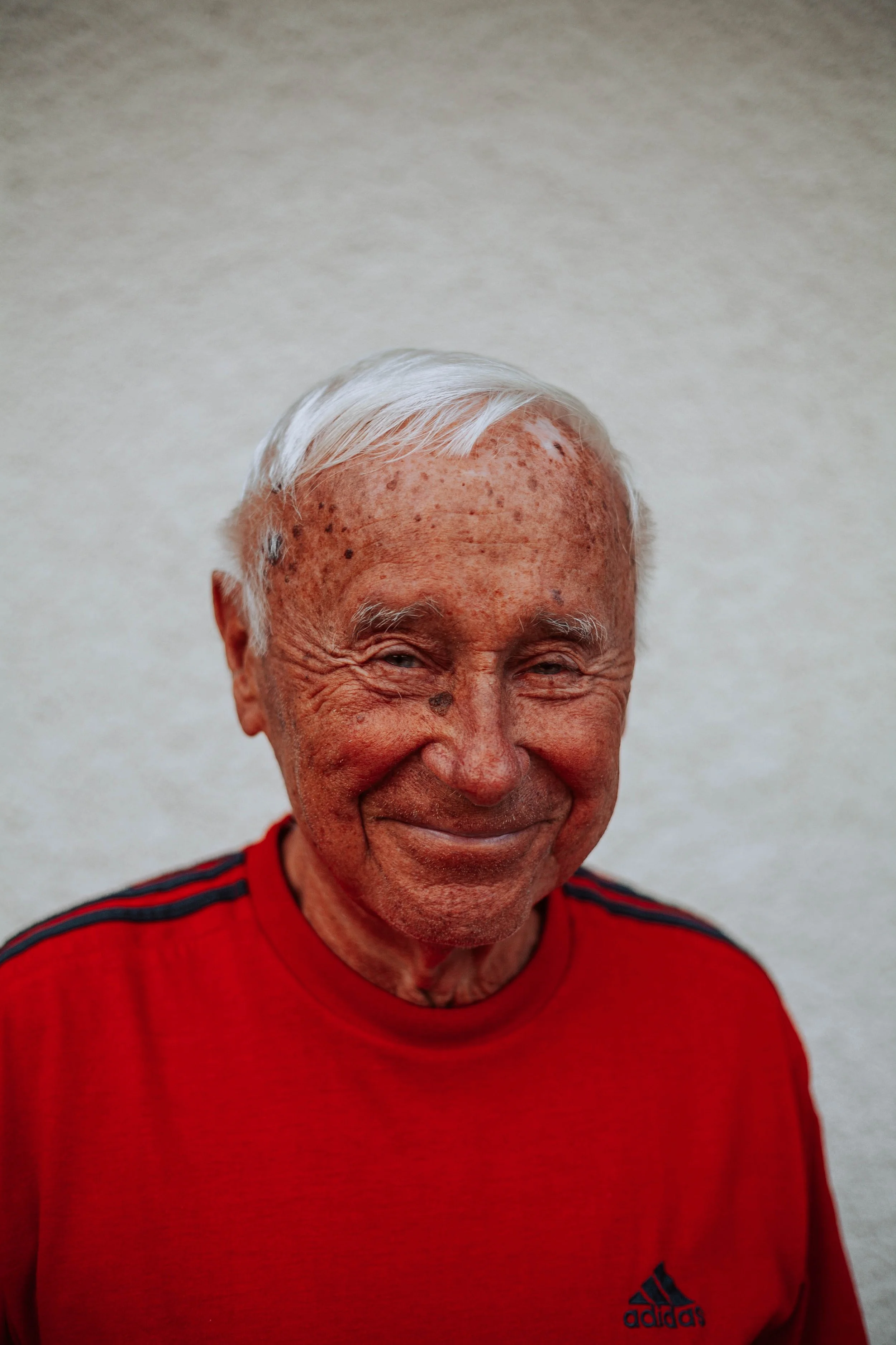 Close-up portrait of an elderly man with white hair, nice smile, and freckles, wearing a red Adidas shirt, standing against a plain light background.
