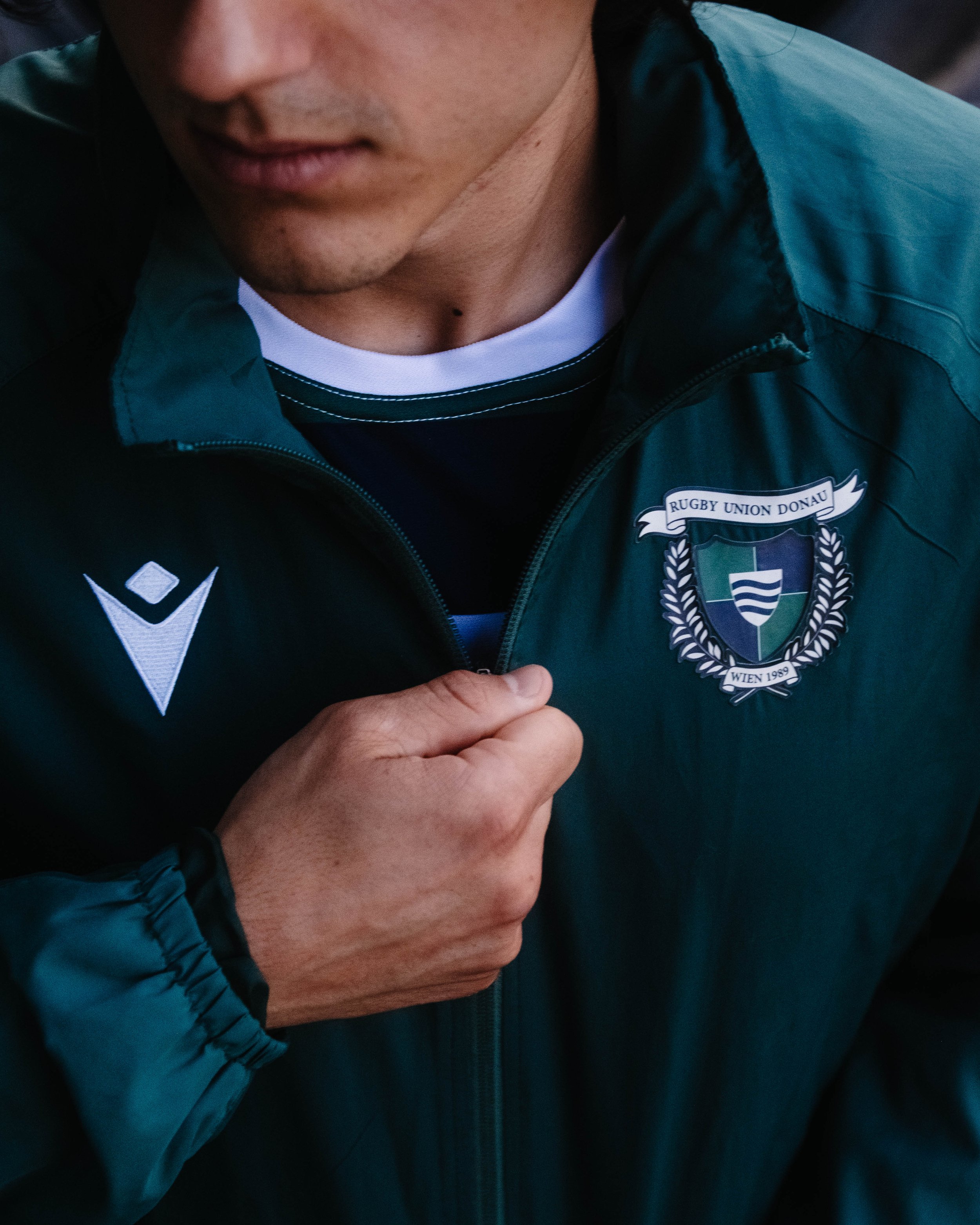 A person wearing a green sports jacket with a crest that reads 'Rugby Union Donau' and a black and white shirt underneath, zipping up the jacket.