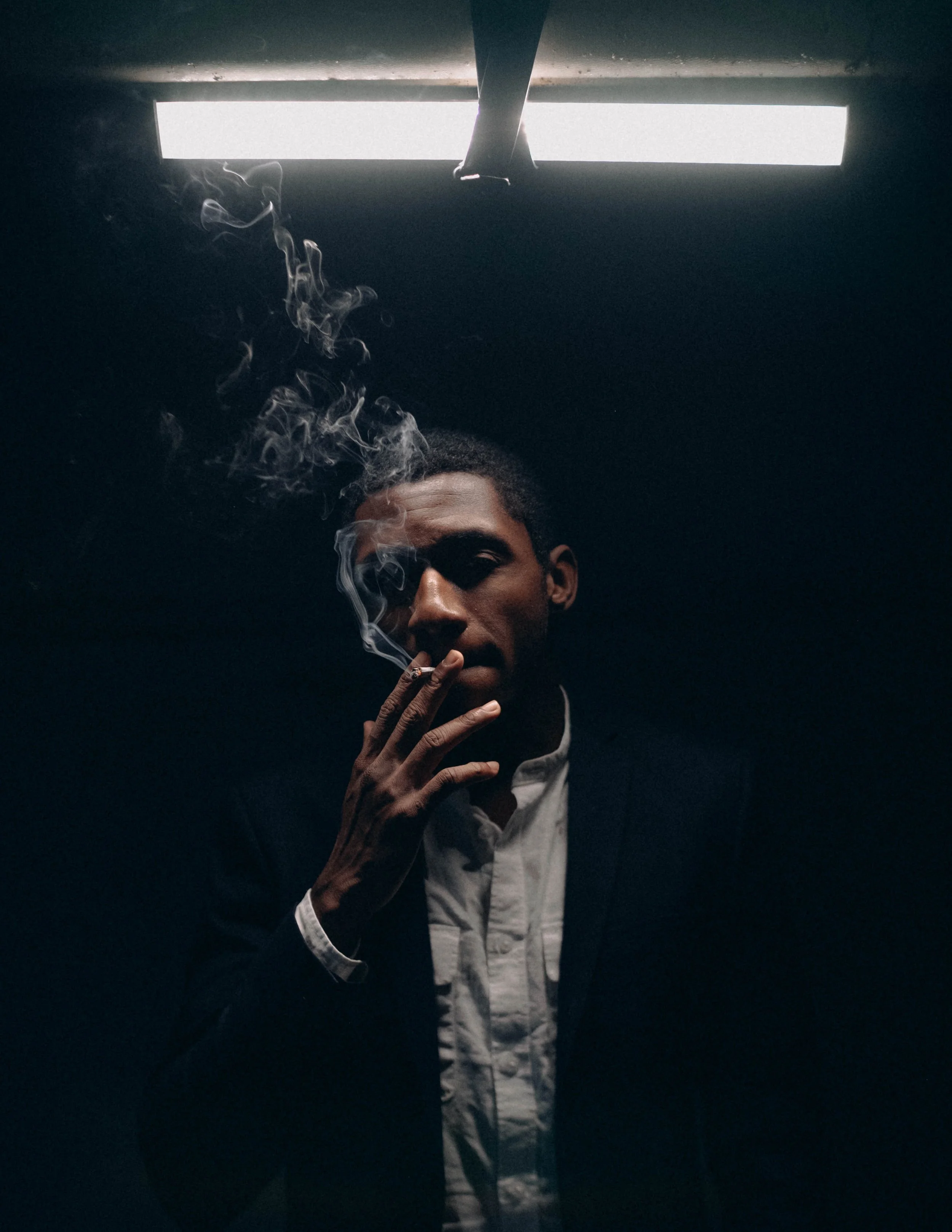 A man in a dark setting, smoking a cigarette with smoke rising, illuminated by a bright overhead light.