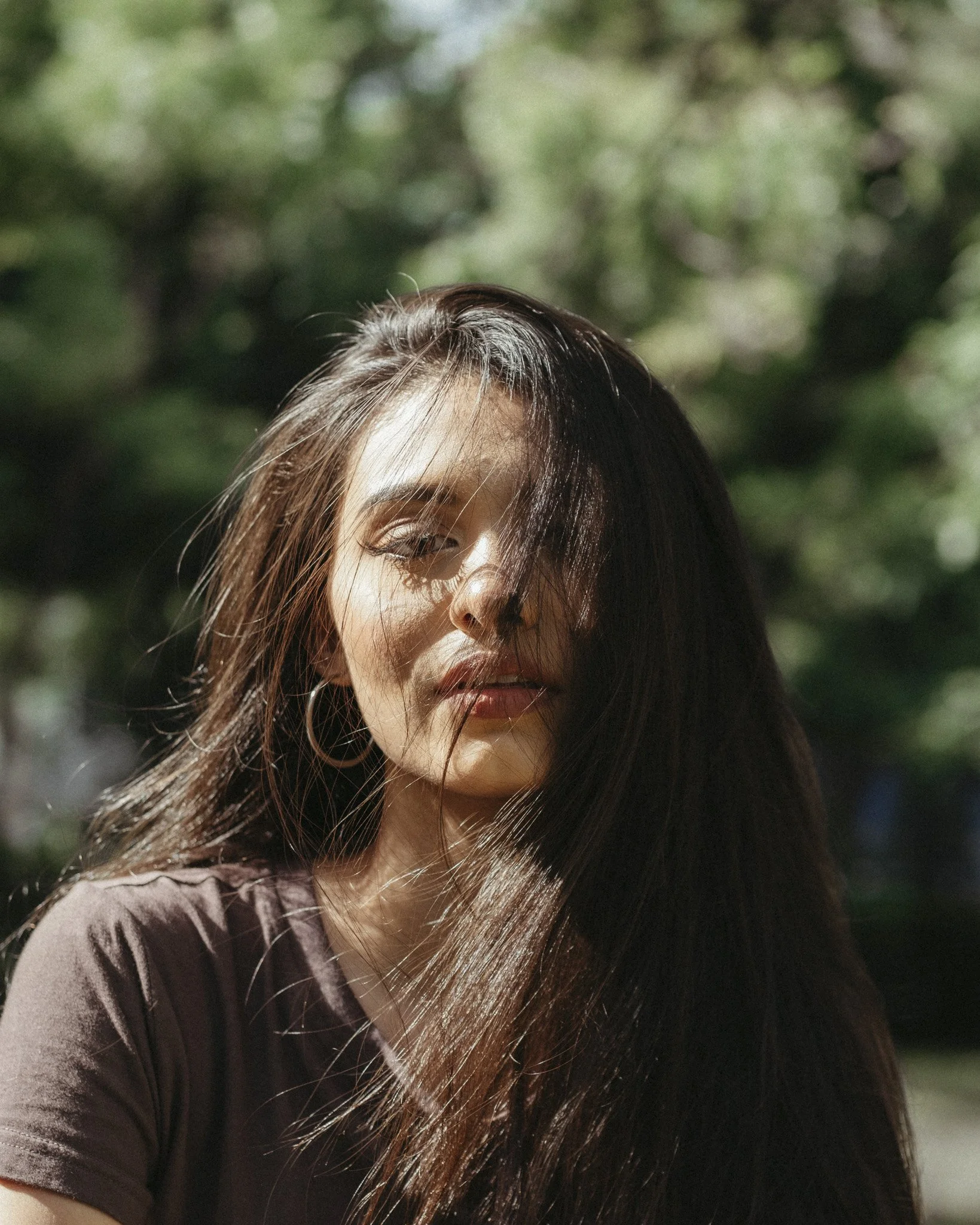 A woman with long brown hair outdoors with sunlight on her face, partially covered by her hair, with a blurred green background.