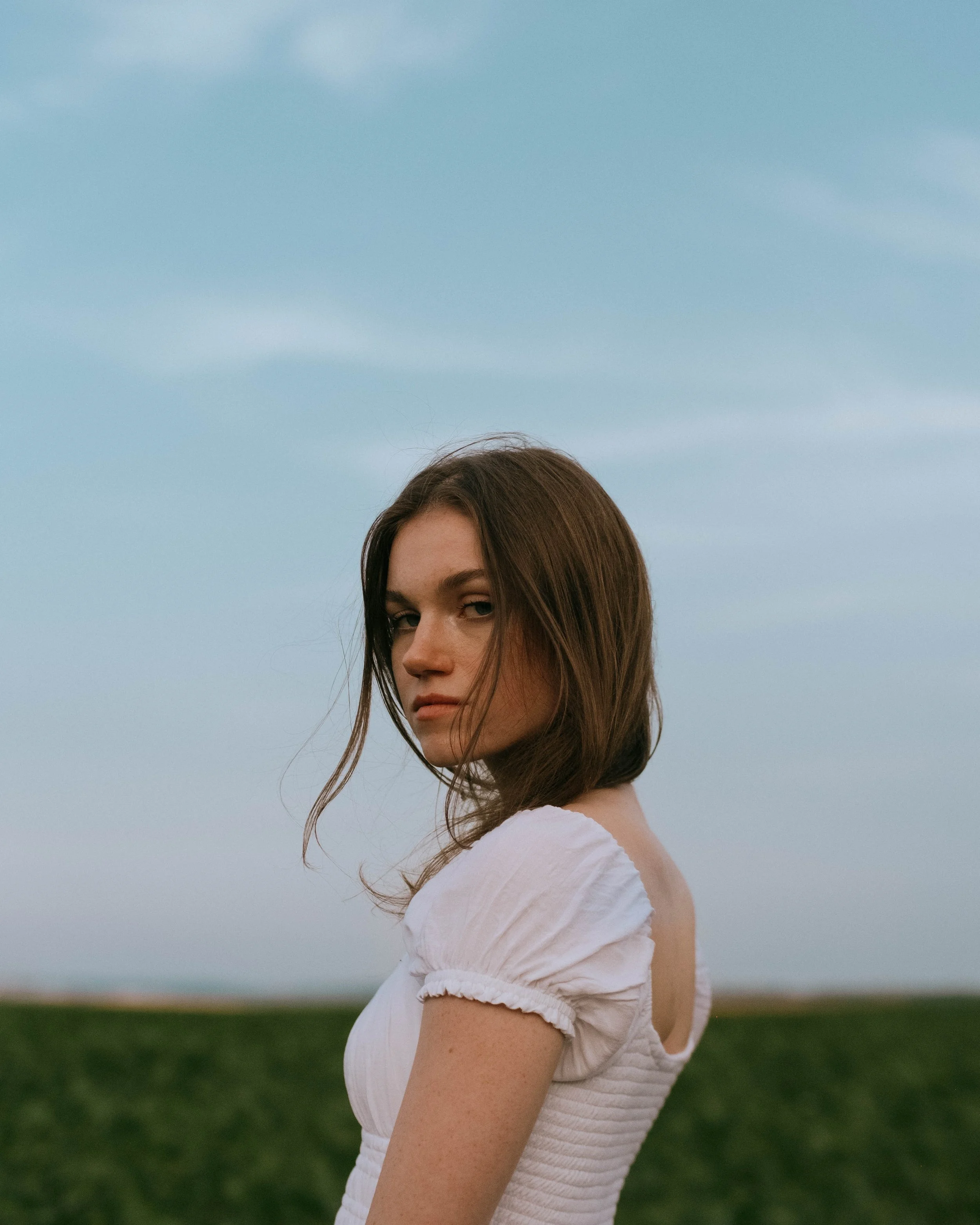 A young woman with brown hair in a white dress, standing outdoors against a landscape with a cloudy sky and green grass.