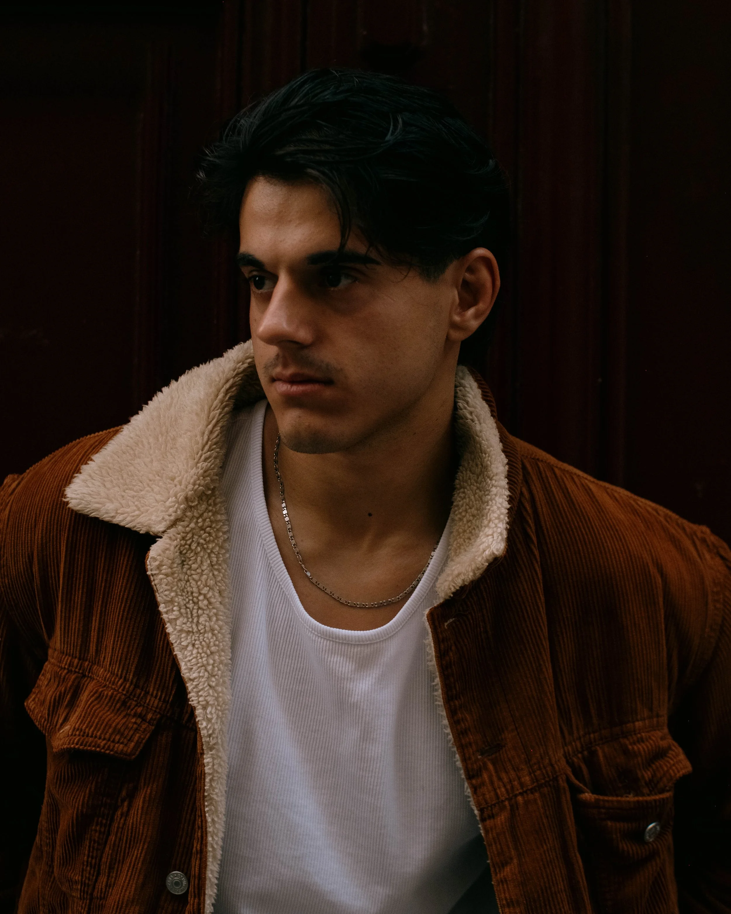 A young man with dark hair, wearing a white t-shirt and a brown corduroy jacket with a shearling collar, looking to the side against a dark wooden background.