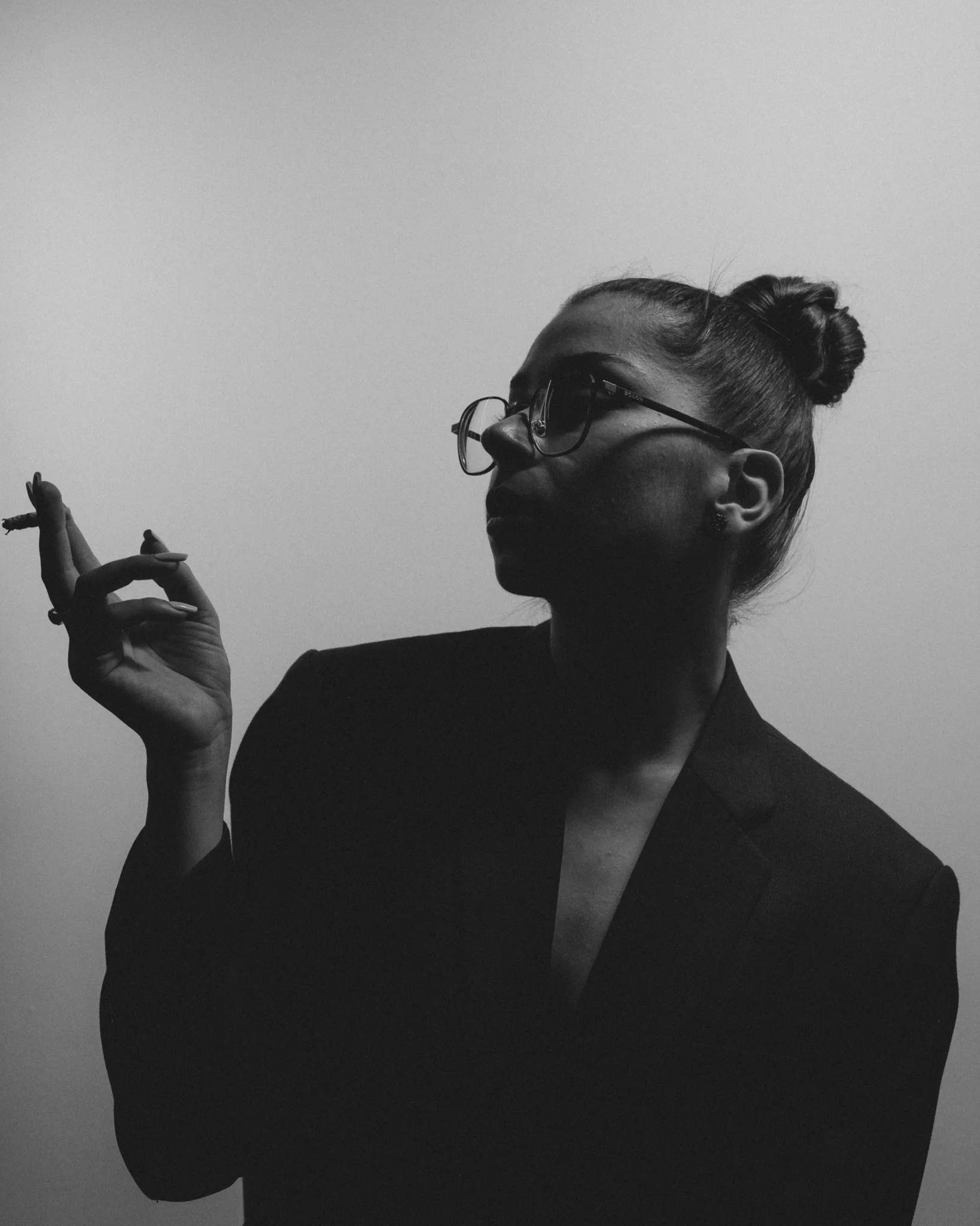 A black and white photo of a woman with glasses and her hair in a bun, holding a cigarette in her right hand, wearing a dark blazer, against a plain light background.