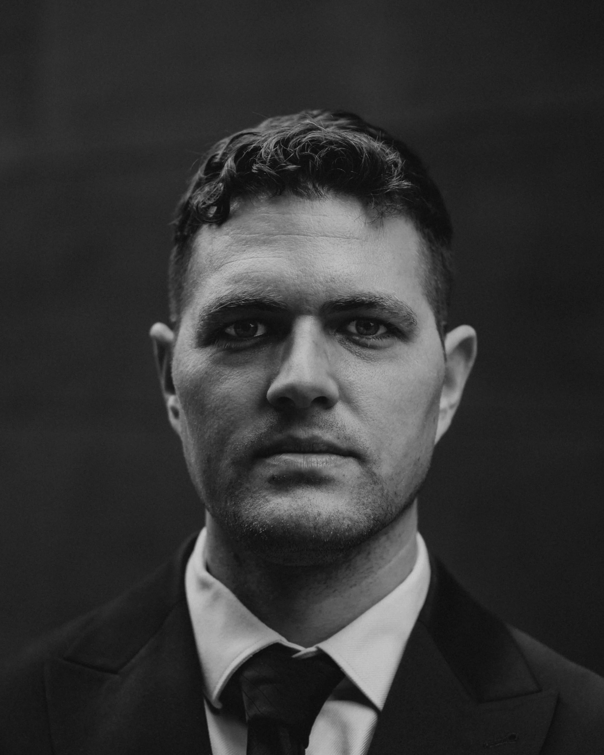A black and white portrait of a man wearing a suit, with a serious expression, facing directly towards the camera.