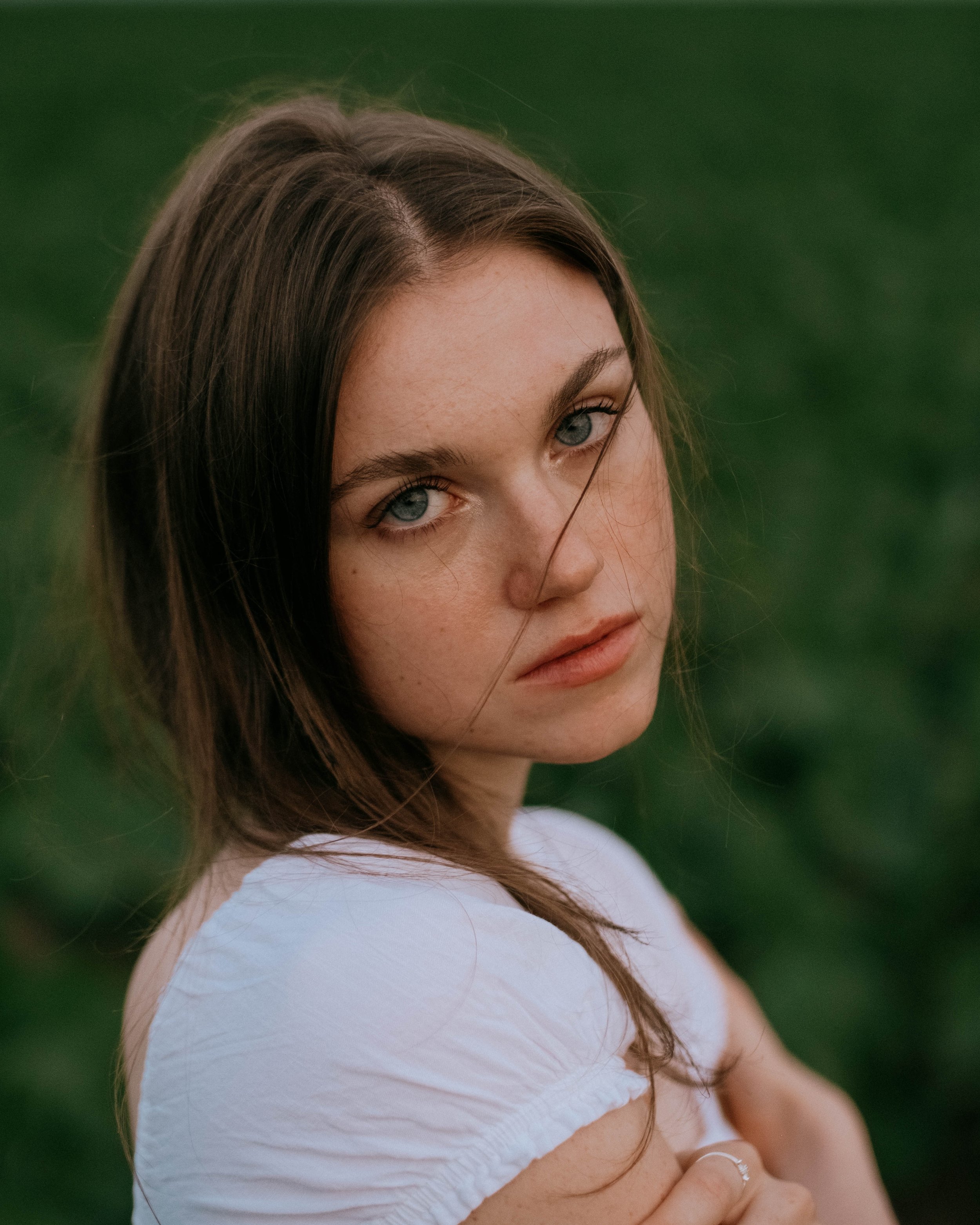 A young woman with long brown hair and light blue eyes looking directly at the camera, in an outdoor setting with a blurred green background.