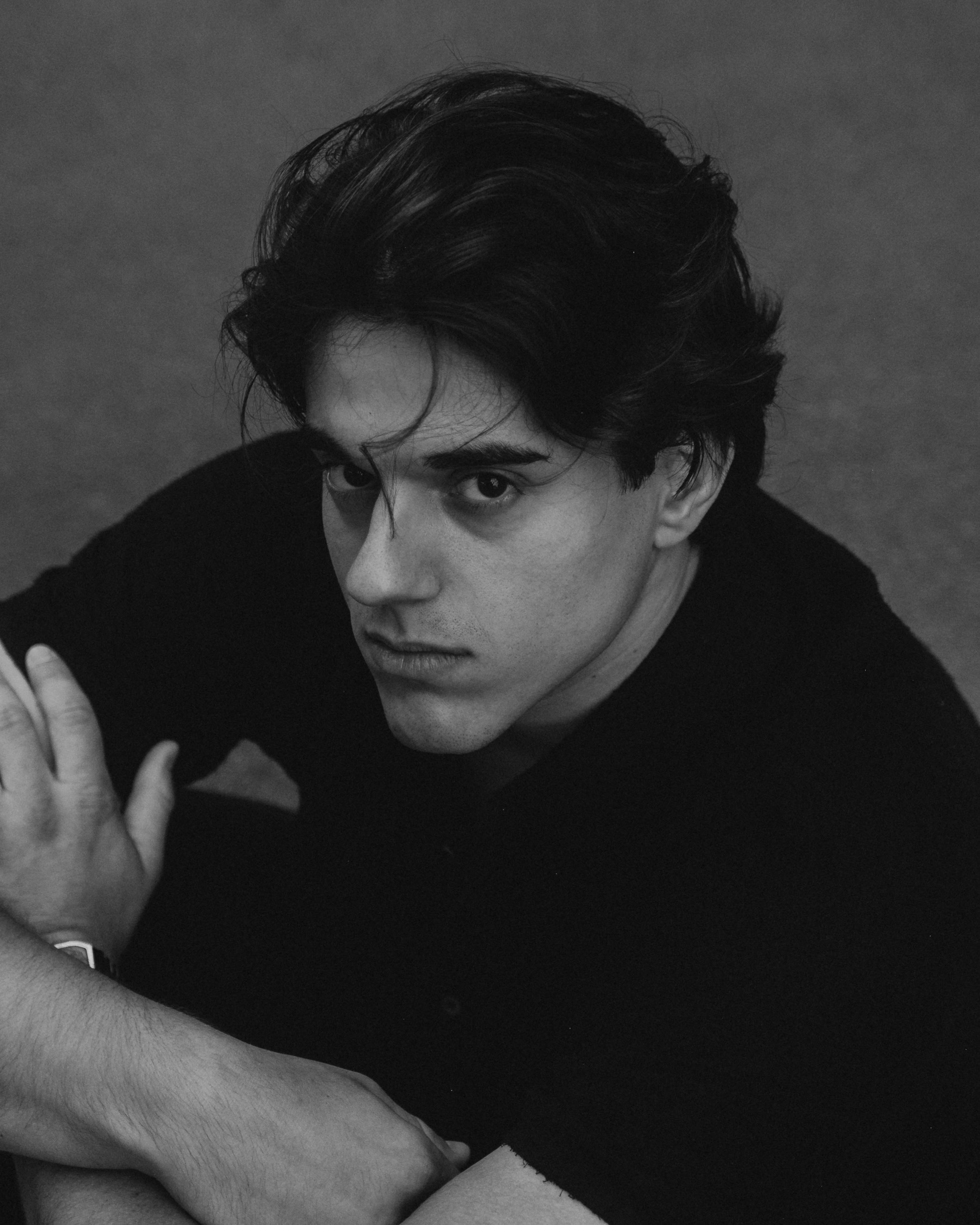 Black and white portrait of a young man with wavy hair, looking up at the camera, arms crossed, wearing a dark shirt.