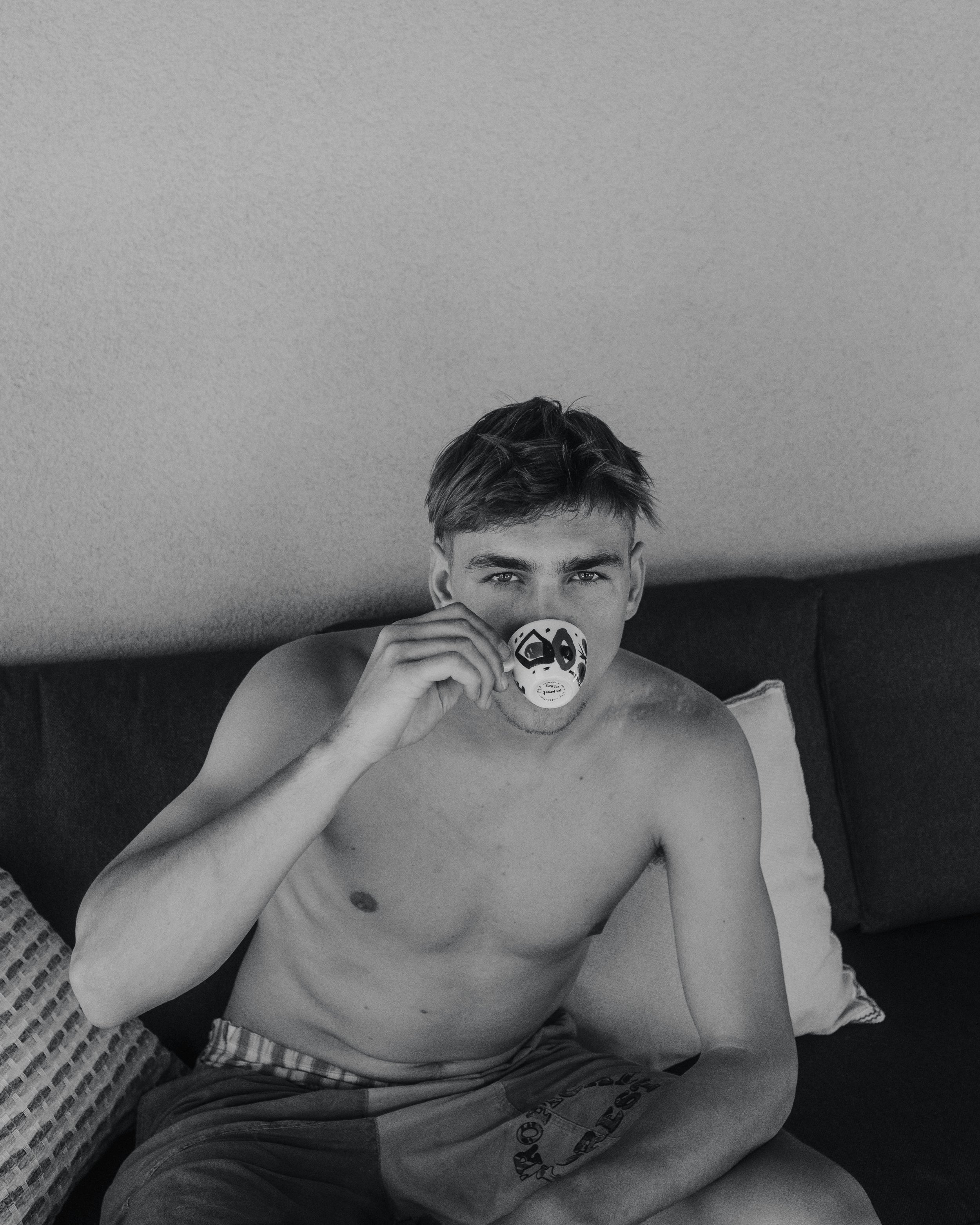 A shirtless young man with short hair sitting on a couch, holding a mask to his face, in a room with a light-colored wall.