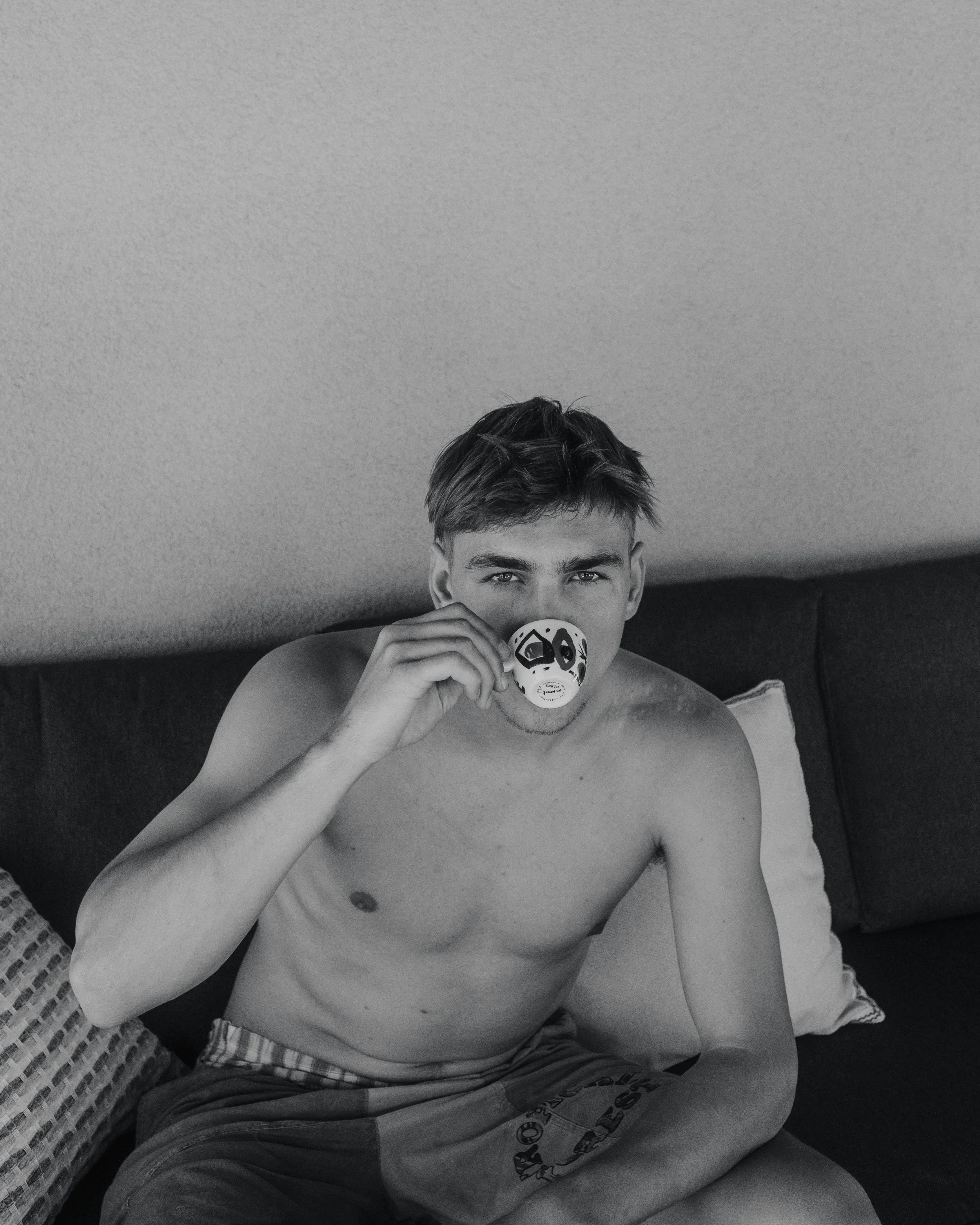 A shirtless young man with short hair sitting on a couch, holding a mask to his face, in a room with a light-colored wall.