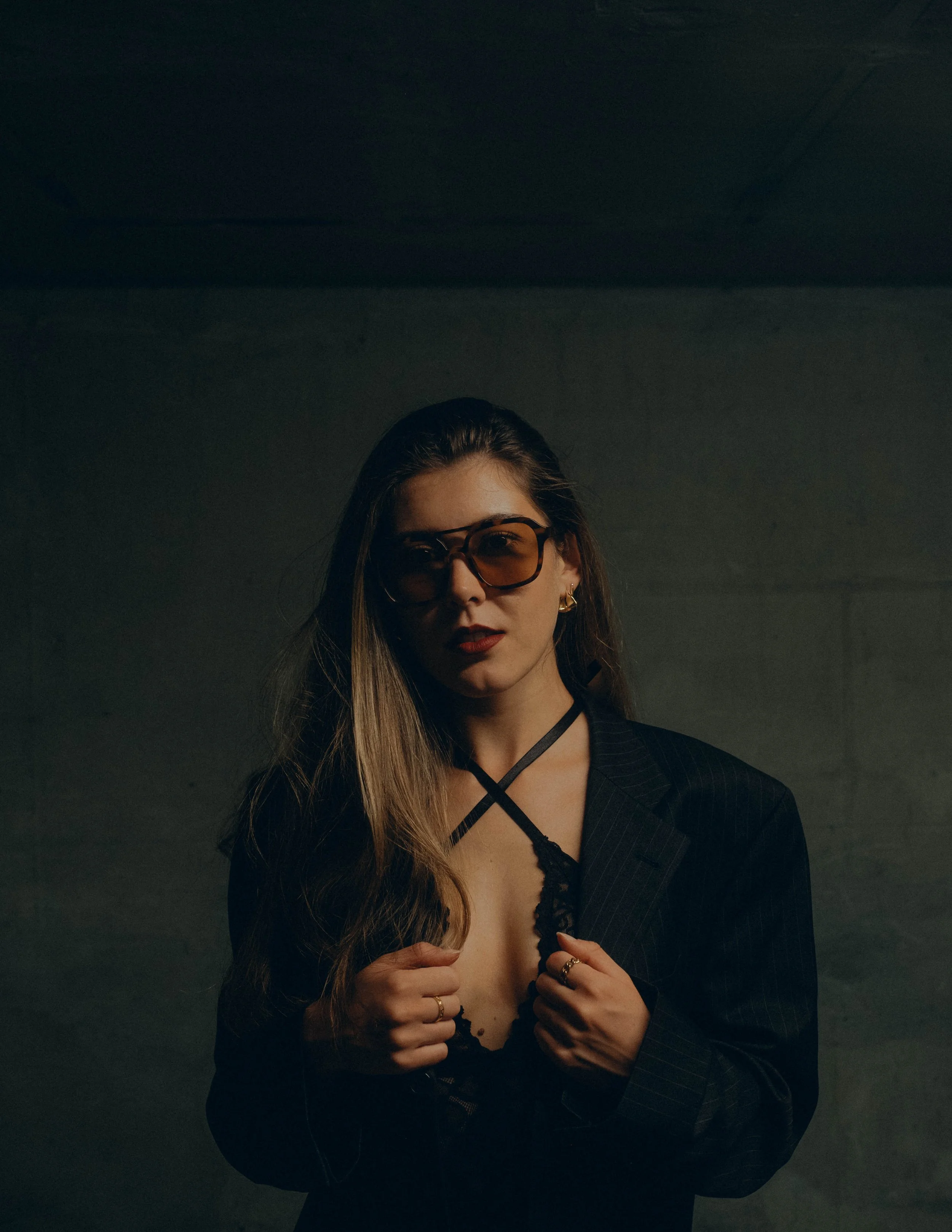 Portrait of a woman with long hair wearing sunglasses, a dark blazer, and lace clothing, standing against a dark background.