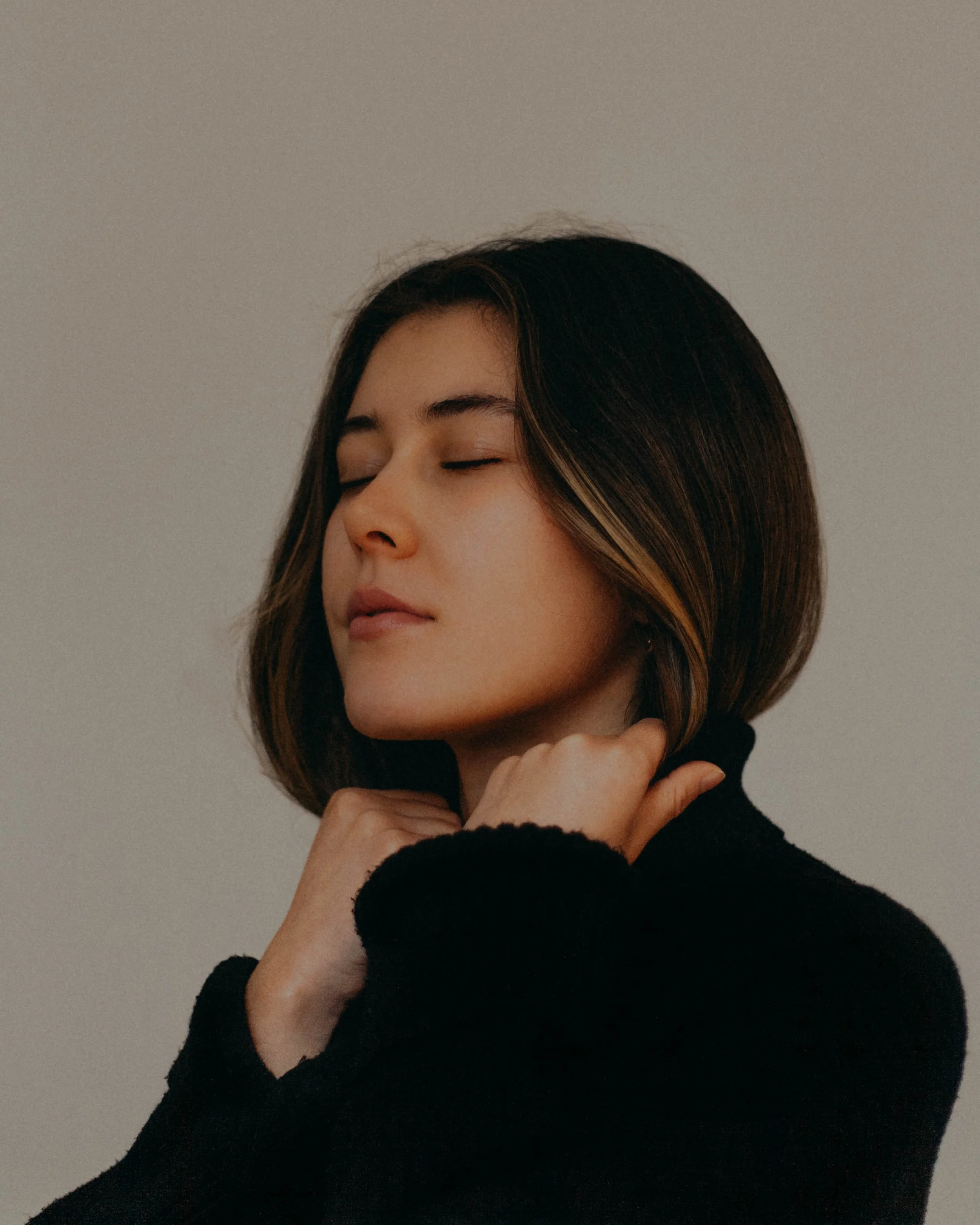 A woman with short dark hair, eyes closed, wearing a black turtleneck, touching her neck, against a plain background.