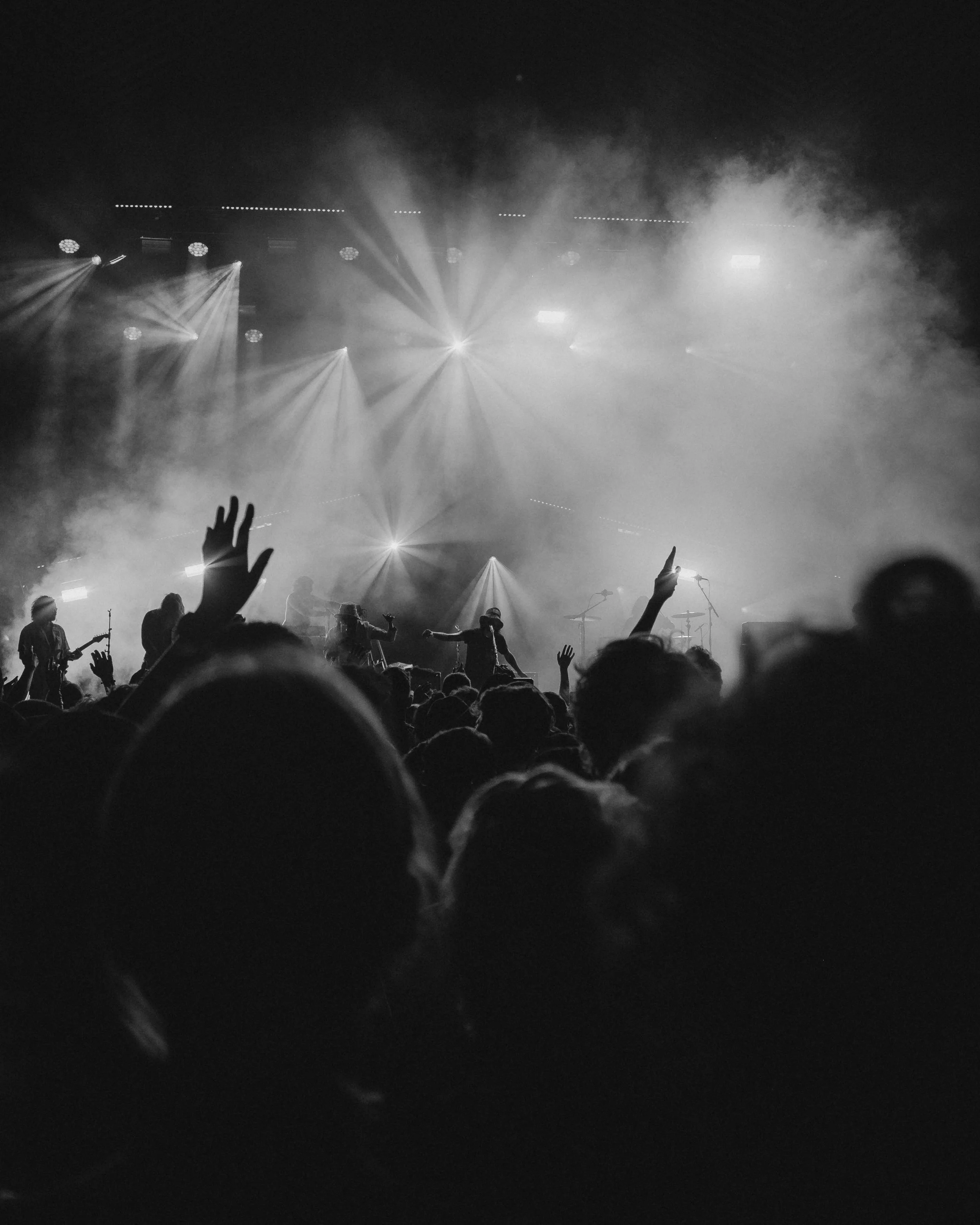 Black and white photo of a concert with a crowd, raised hands, and stage lights shining through smoke.