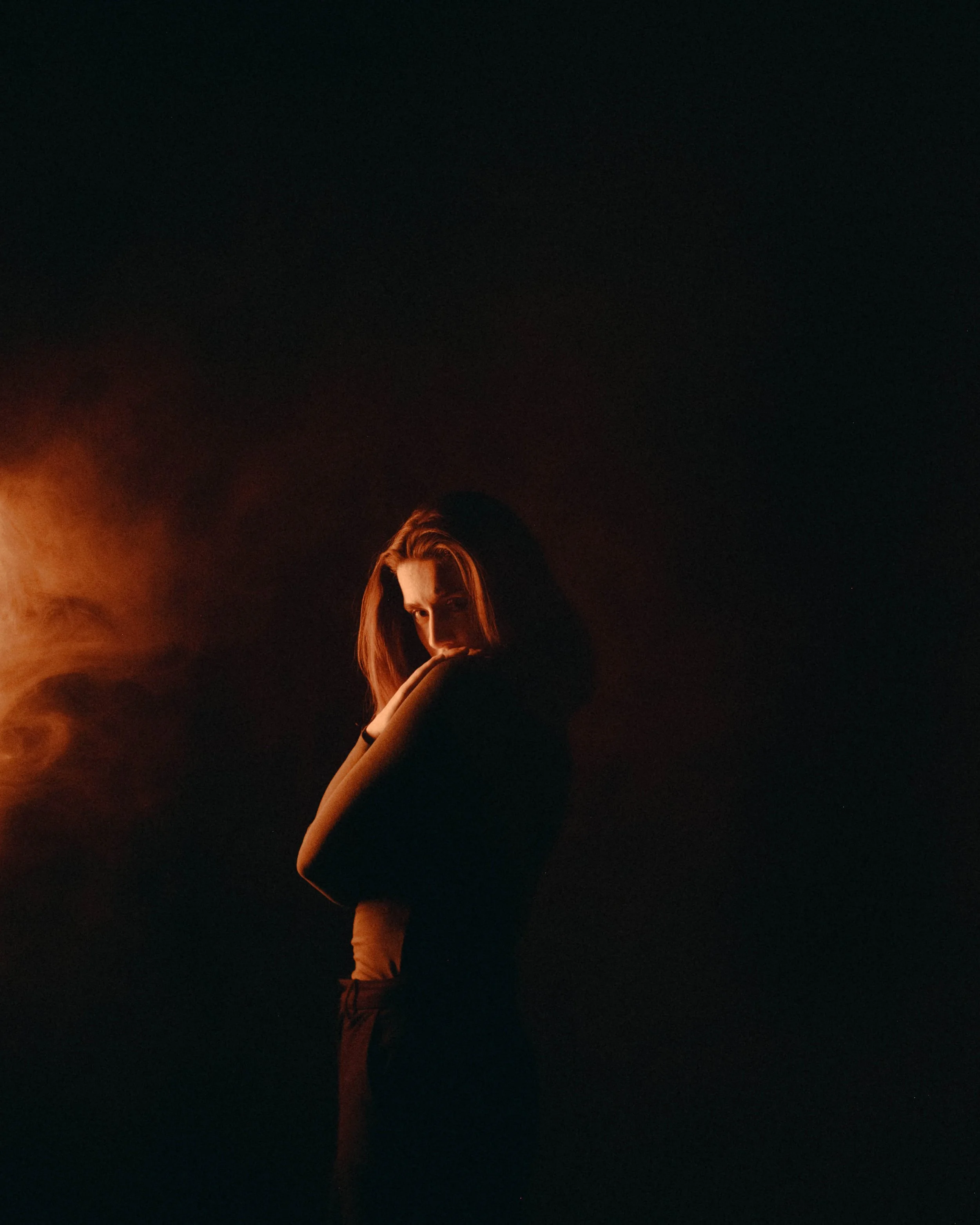 A woman with red hair standing in darkness with a warm light illuminating her face and arm, looking pensively downward with her arms crossed.