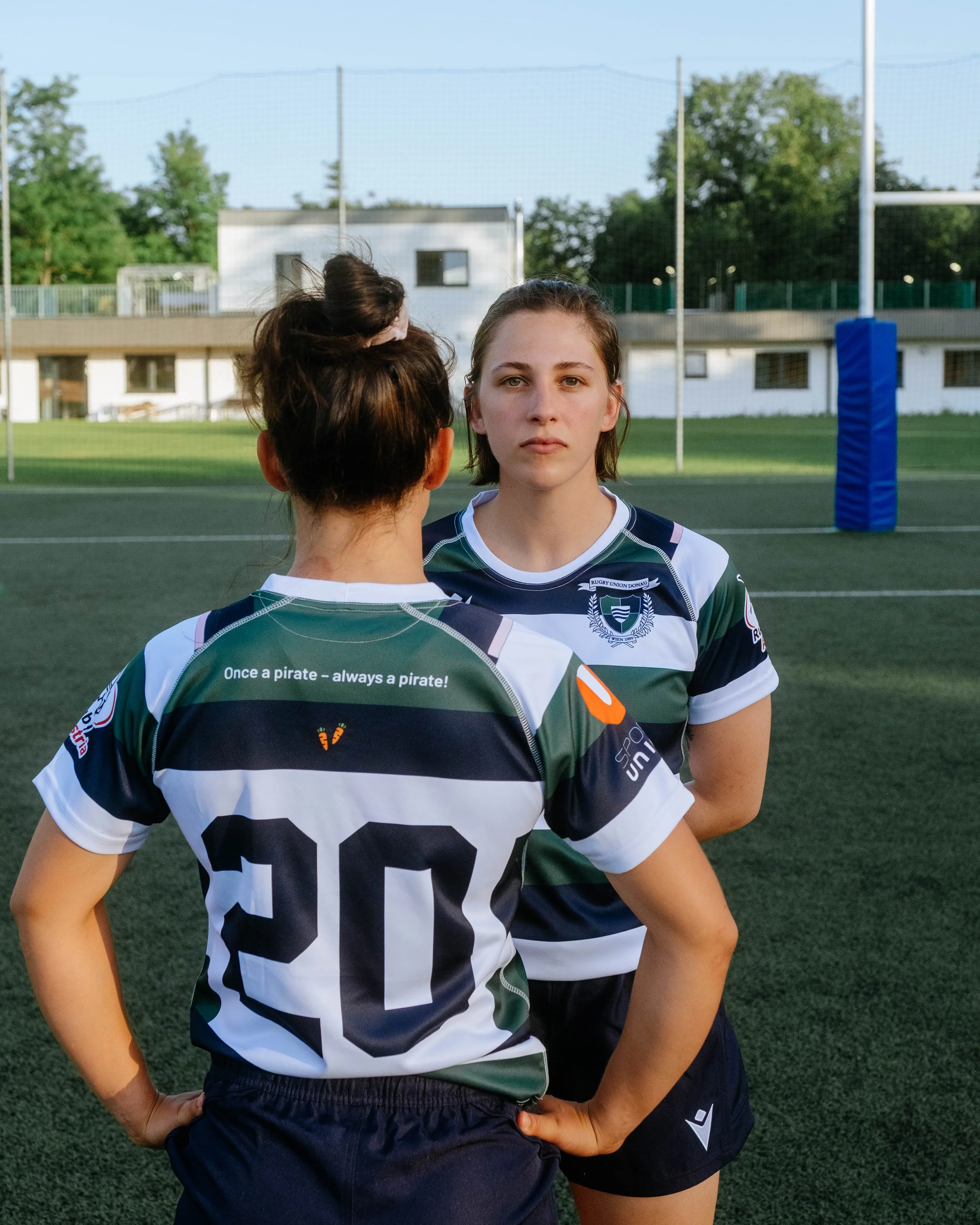 Two female rugby players wearing green, white, and black jerseys standing on a rugby field, facing each other with serious expressions.