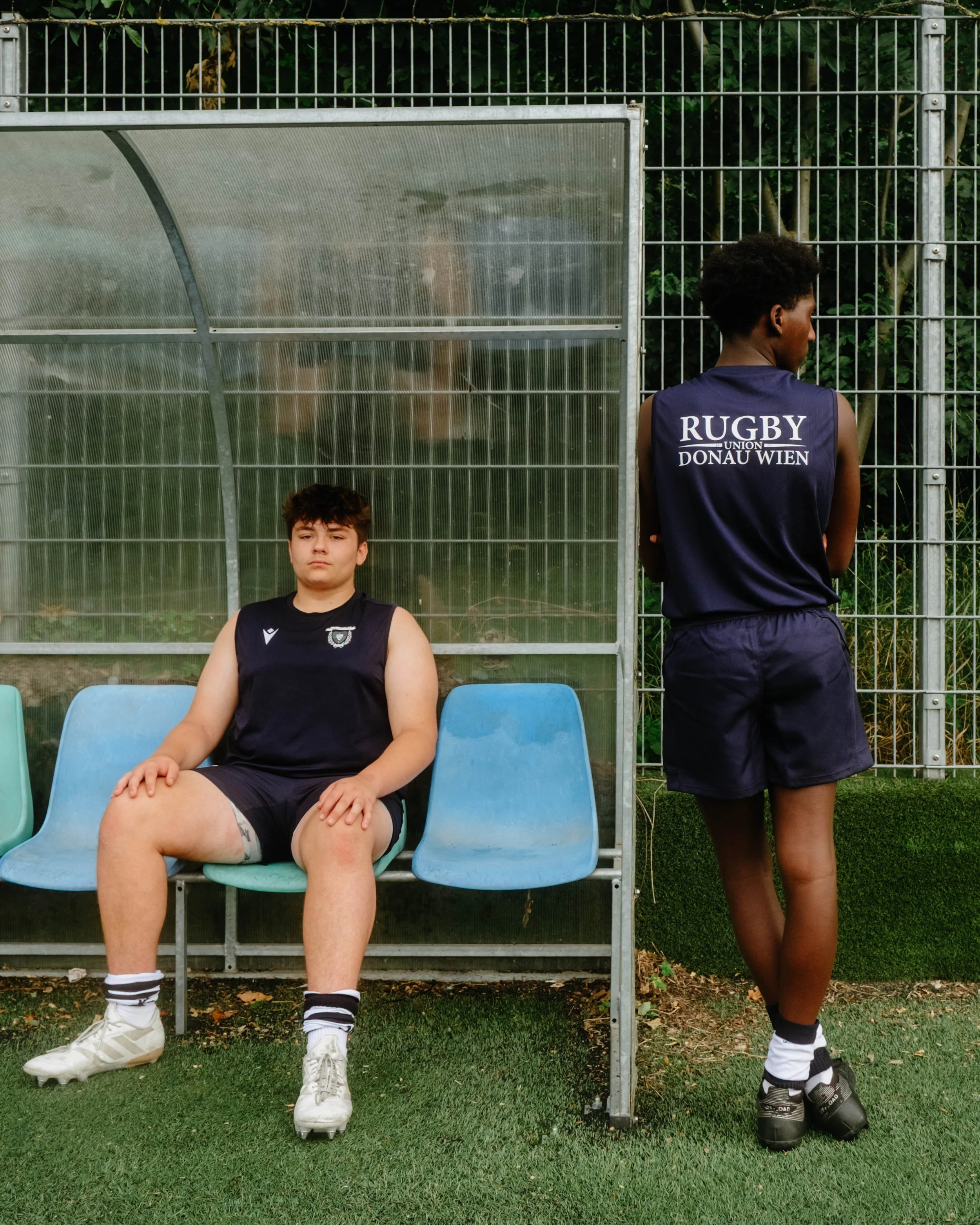Two young rugby players in uniform on a sideline bench, one sitting and one standing, behind a metal fence at a sports field.