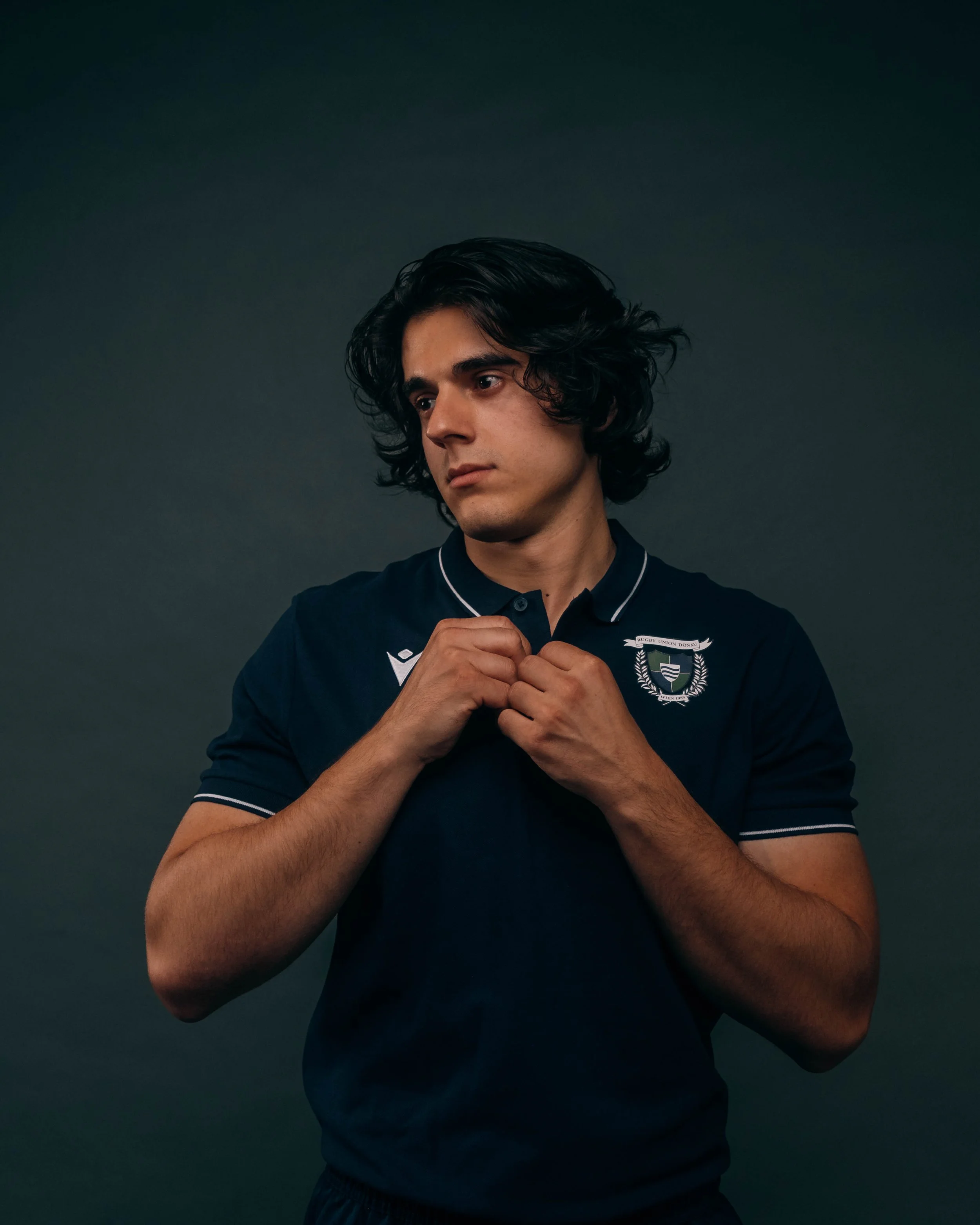 Young man with dark, wavy hair adjusting a navy blue sports jersey with a crest and white accents against a dark background.