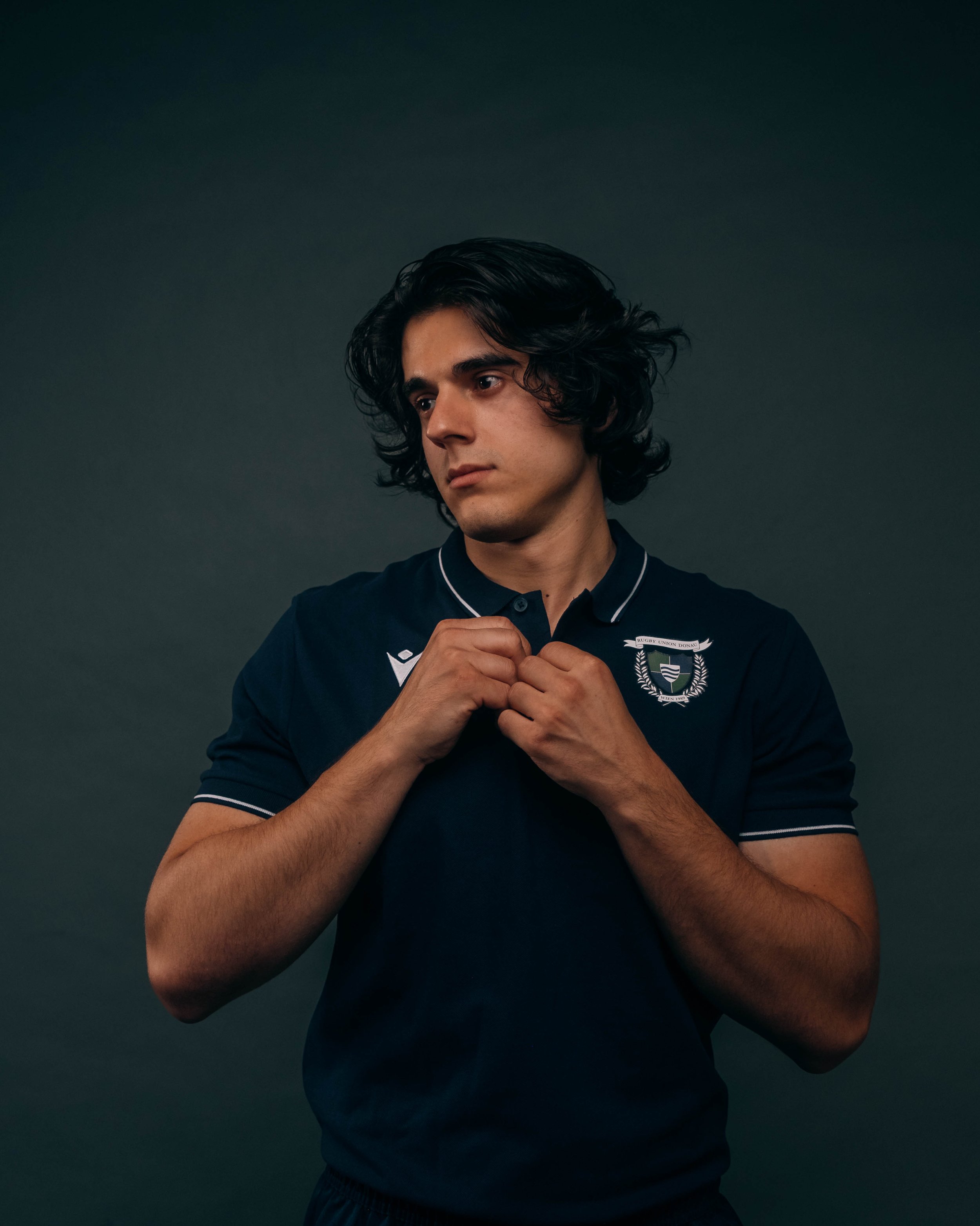 Young man with dark, wavy hair adjusting a navy blue sports jersey with a crest and white accents against a dark background.