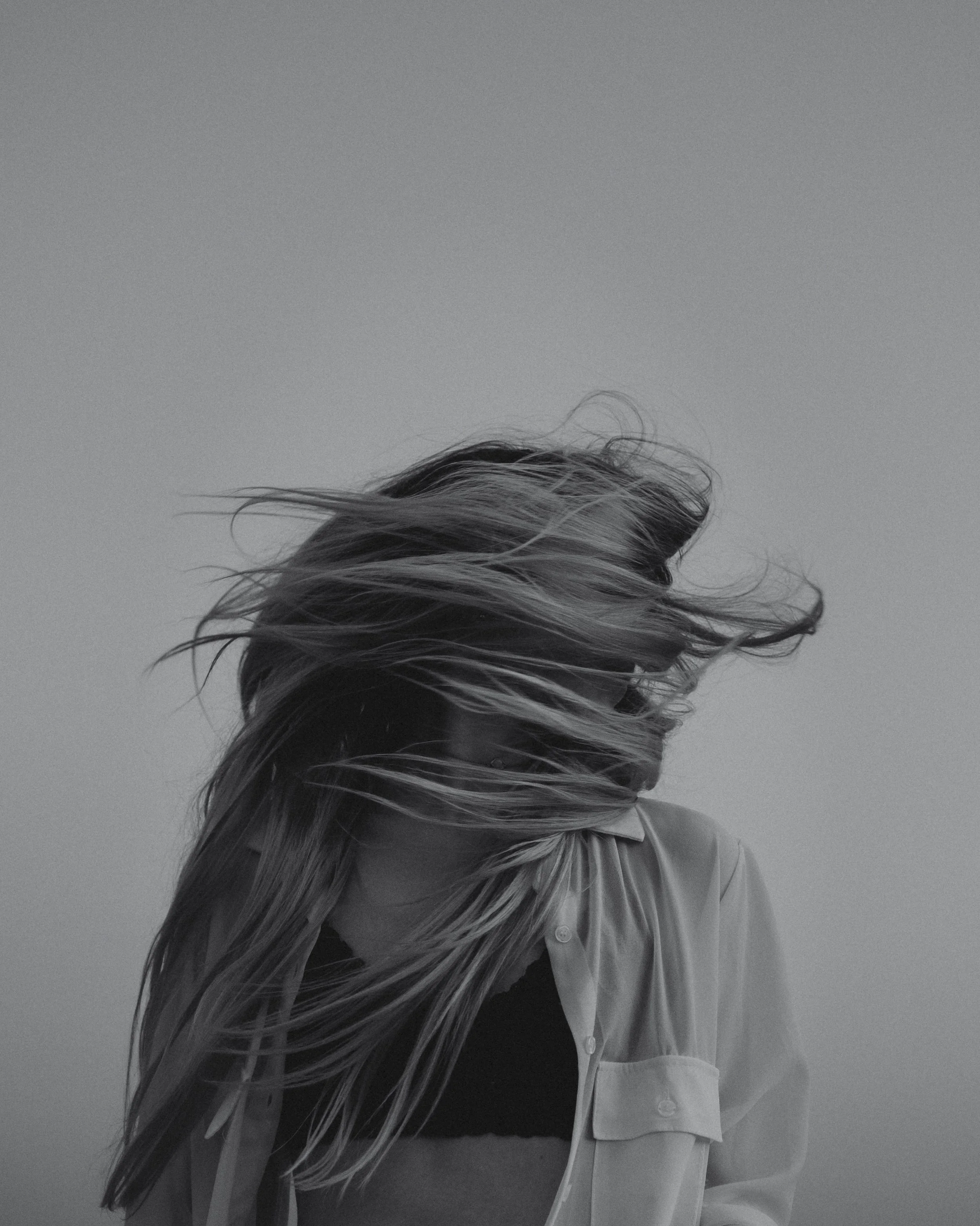 A black and white photo of a woman with long hair, her head tilted downward, hair blowing in the wind, wearing a light-colored shirt over a dark top.
