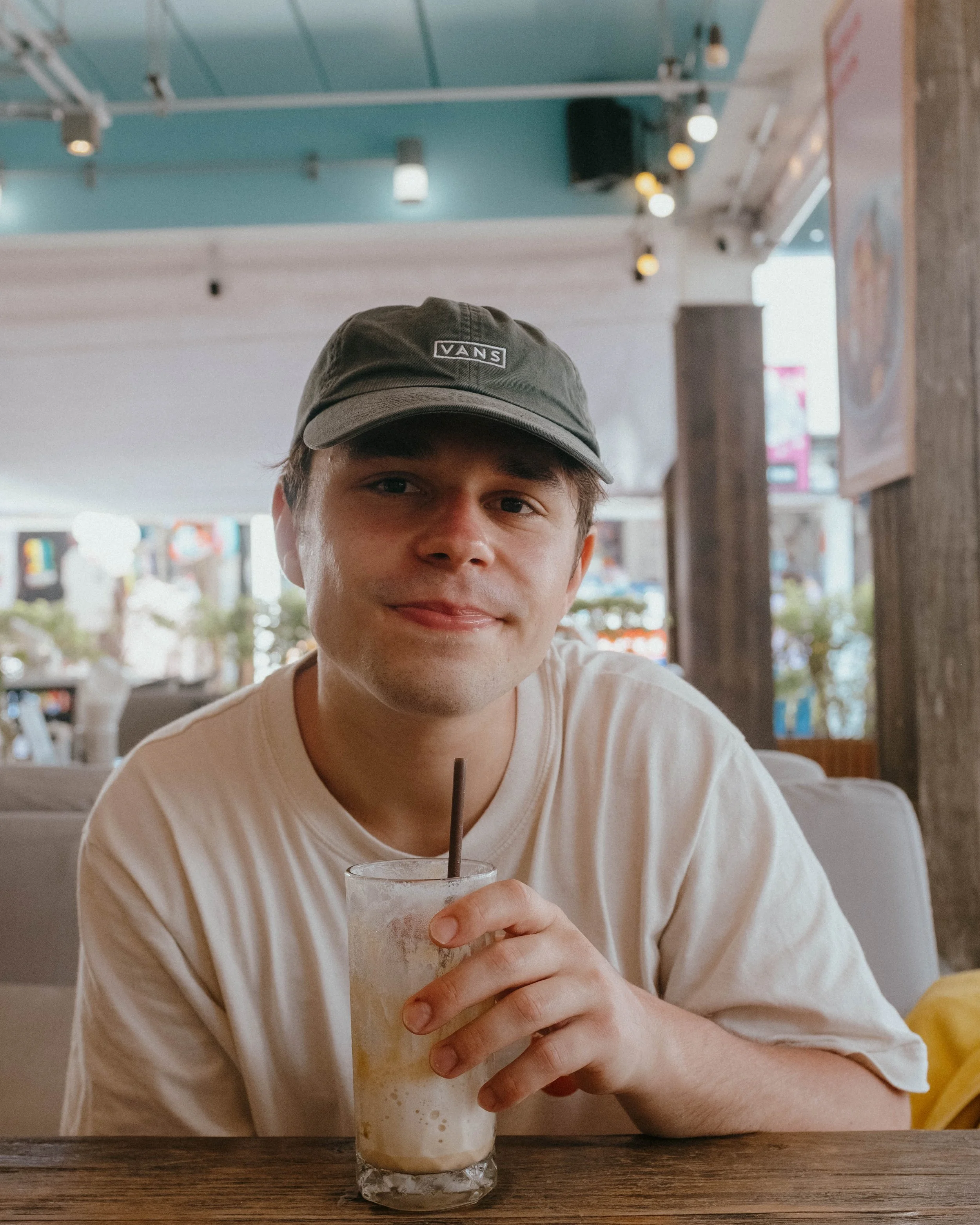 A young man in a gray cap and beige T-shirt sitting at a restaurant table, holding a glass of iced drink with a straw, smiling at the camera.