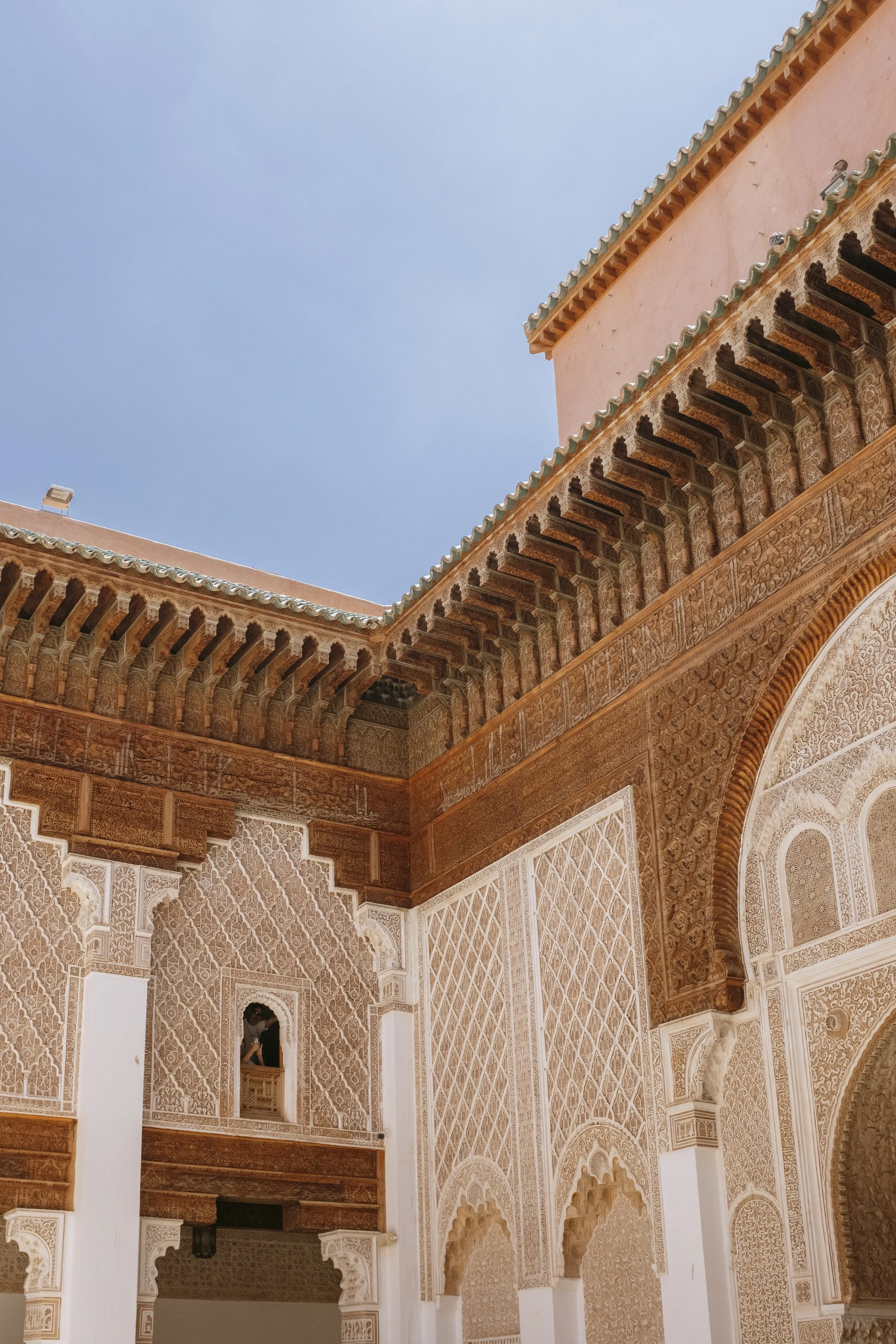 Interior view of a historic building with ornate Moroccan-style white stucco carvings and archways, and a section of the orange exterior wall under a clear blue sky.