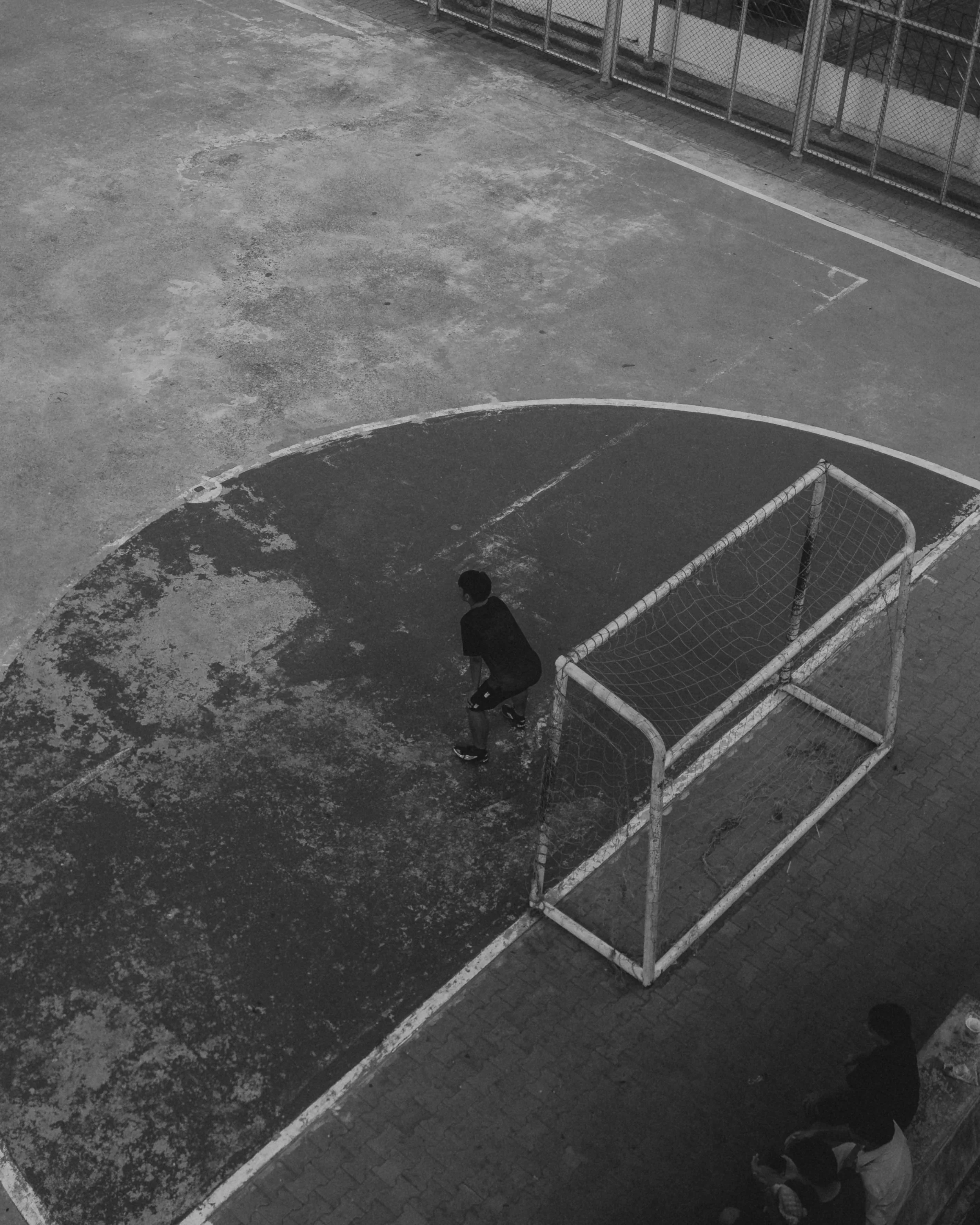 A black and white photo of a boy standing near a small goal on a worn outdoor sports court, with another person sitting on a bench nearby.