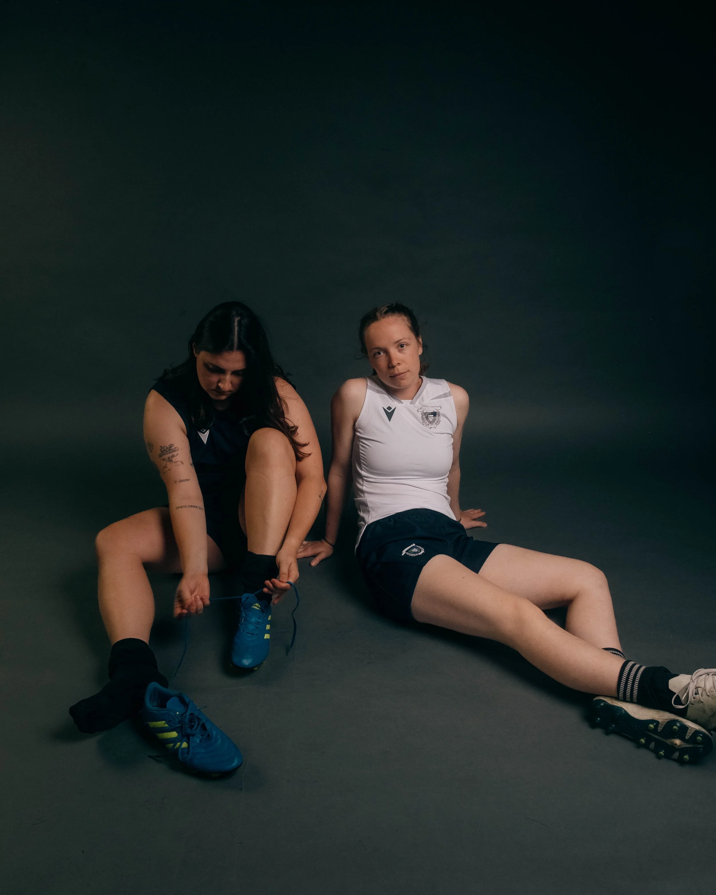Two female athletes sitting on the floor, one adjusting her athletic shoe, both wearing sports uniforms, with dark background.