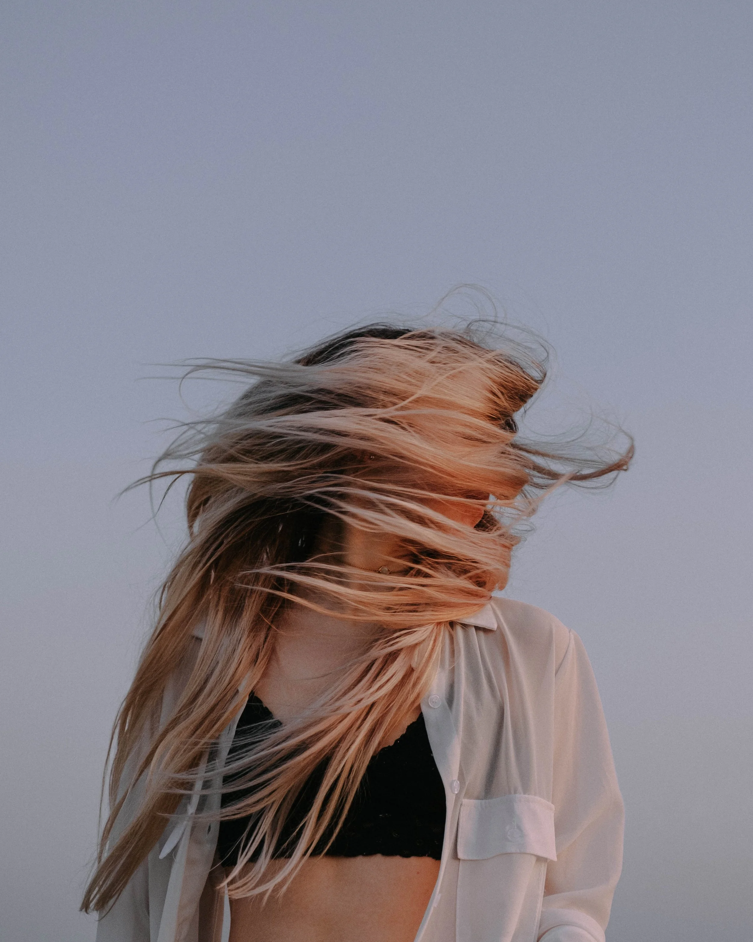 A woman with long, blonde hair, wearing a black crop top and an open beige shirt, standing against a plain sky background with her hair blowing in the wind.