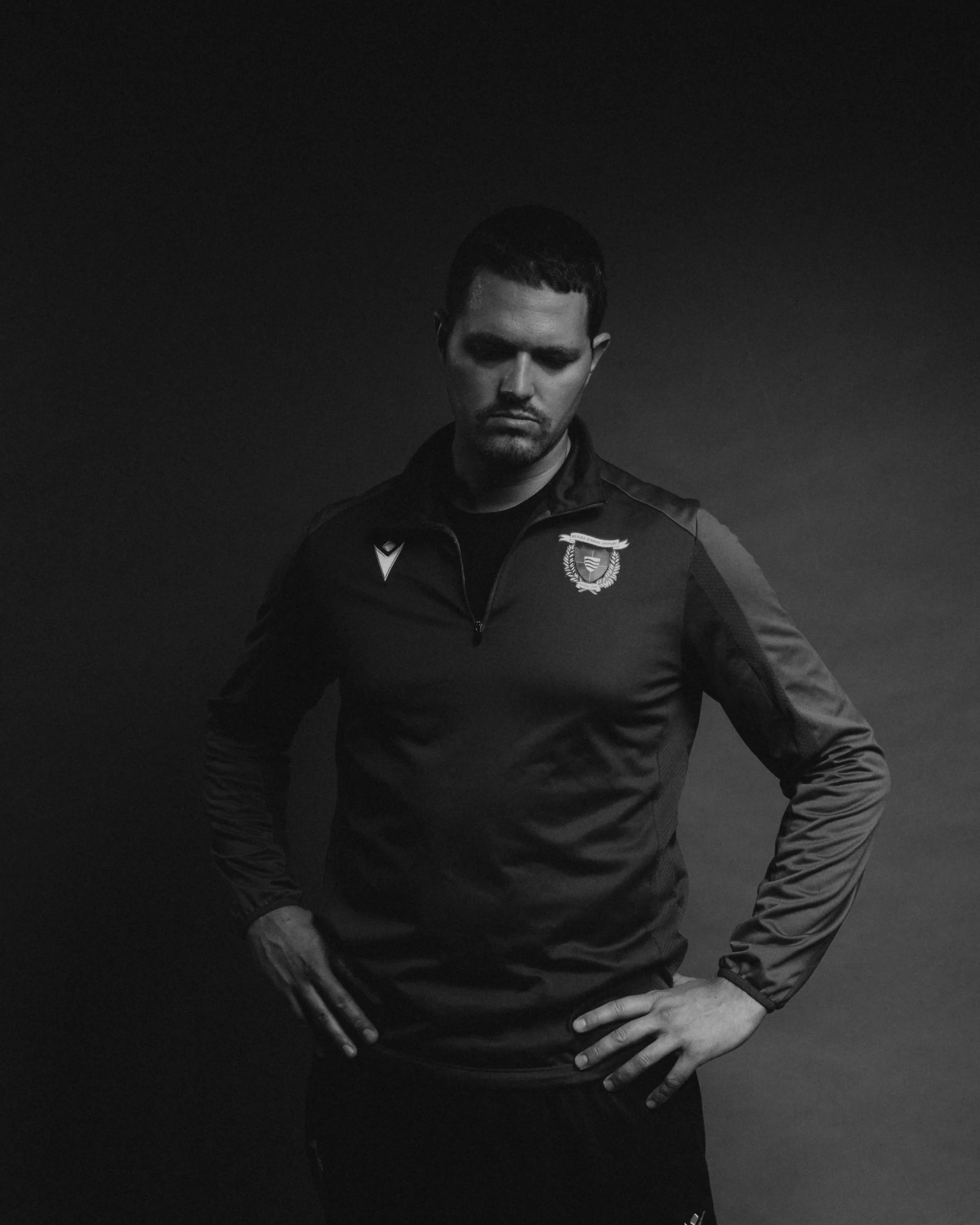 A man in a dark sports jacket standing against a dark background, looking down with hands on hips.