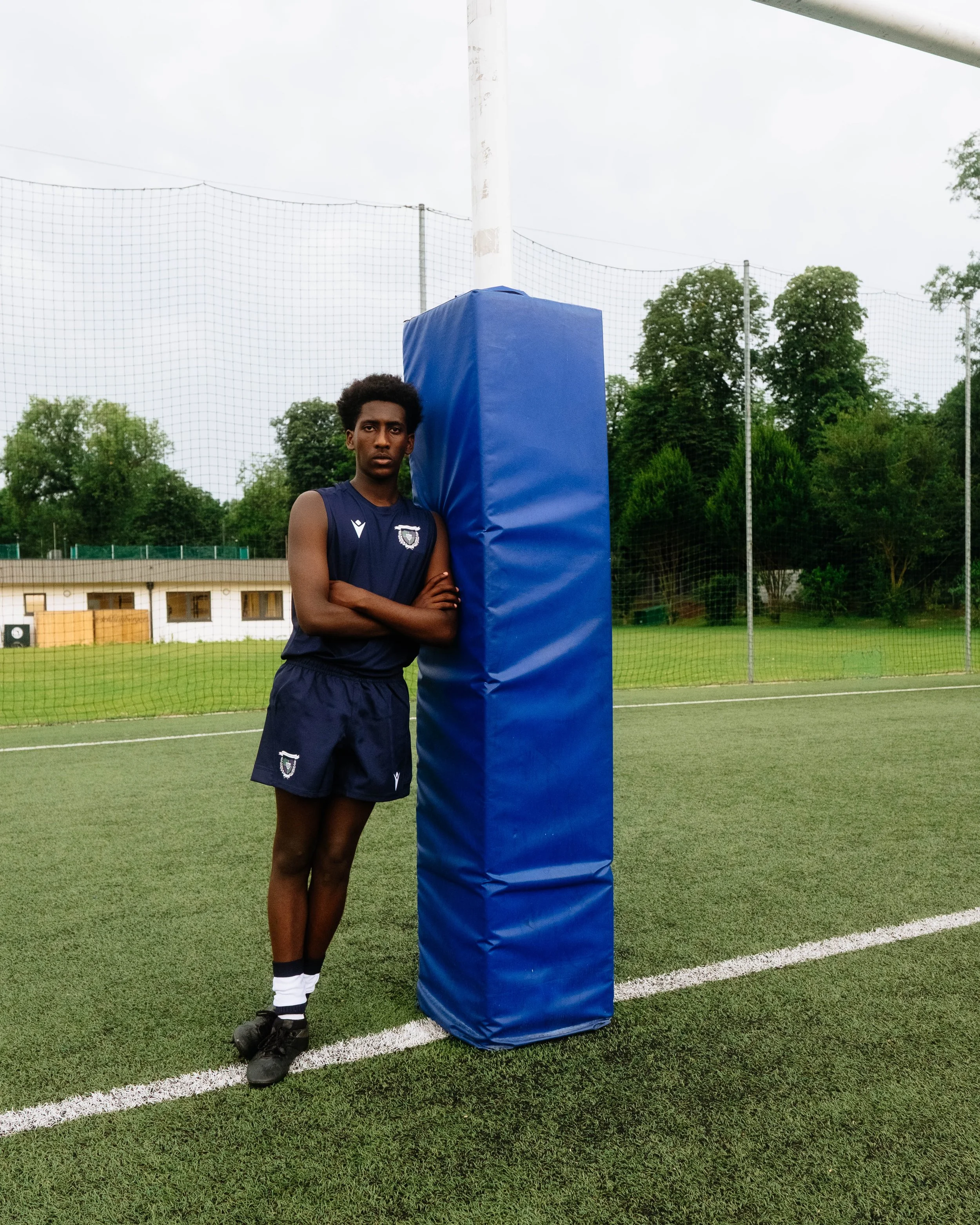 A young man in a sports uniform standing next to a padded post on a sports field, with a netted fence and trees in the background.